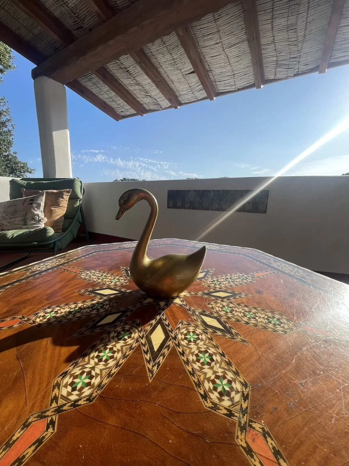 A golden swan figurine on a decorative wooden table with intricate inlay pattern. In the background, there is a cushioned lounge chair with pillows, a white wall, and an outdoor view of blue sky with some clouds and trees under a shaded patio with wooden beams and a thatched roof.