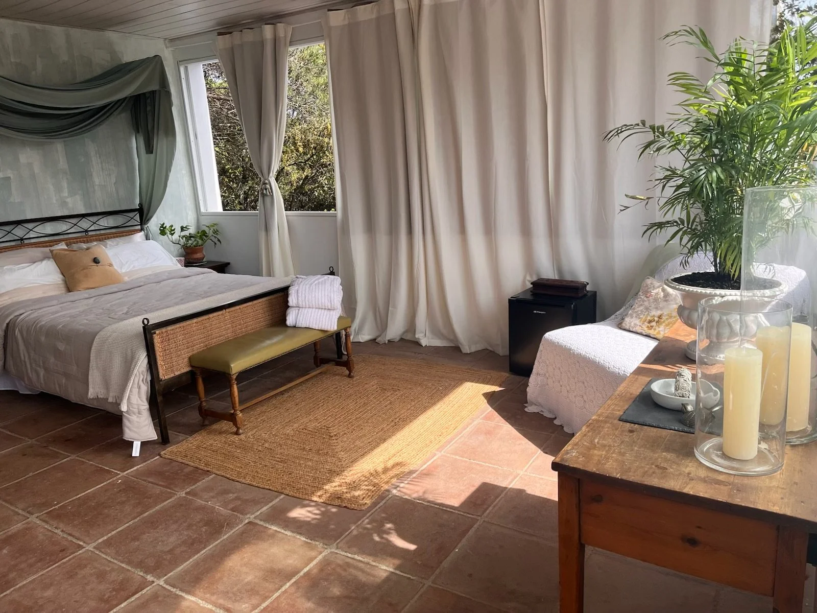 Cozy bedroom with a large bed, nightstand, a bench at the foot of the bed, potted plants, a rug, and a table with candles and decorative items. Sunlight streams through windows with cream curtains.
