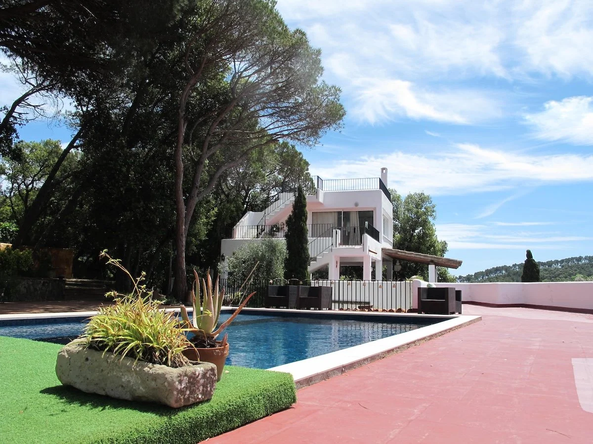 A house with a flat roof and white exterior, surrounded by trees, with an outdoor swimming pool in the foreground. The pool area has red patio flooring and potted plants, and the scene is set on a bright, partly cloudy day.