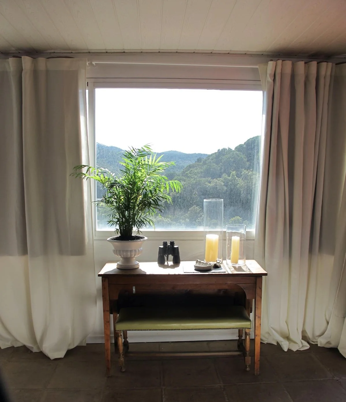 A cozy room with a large window showing a scenic view of green hills and trees. In front of the window, there is a wooden table with potted plant, binoculars, candles, and decorative items. Cream-colored curtains frame the window, and a cushioned stool is placed beneath the table.