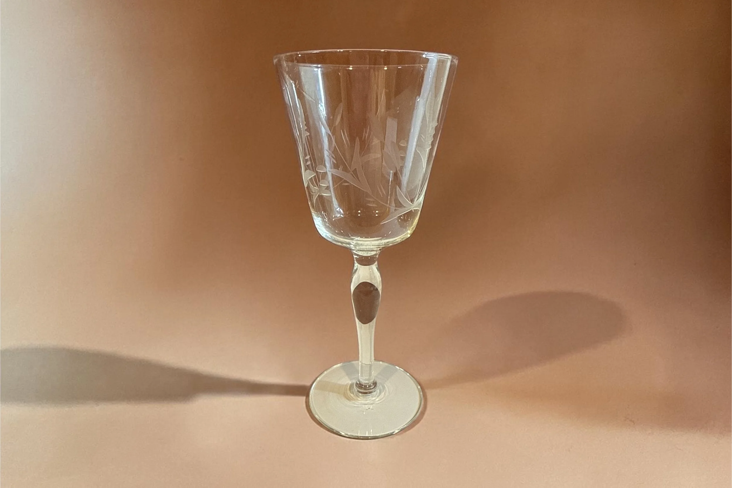A clear glass with an etched floral pattern, standing on a round base, cast a shadow on a neutral background.