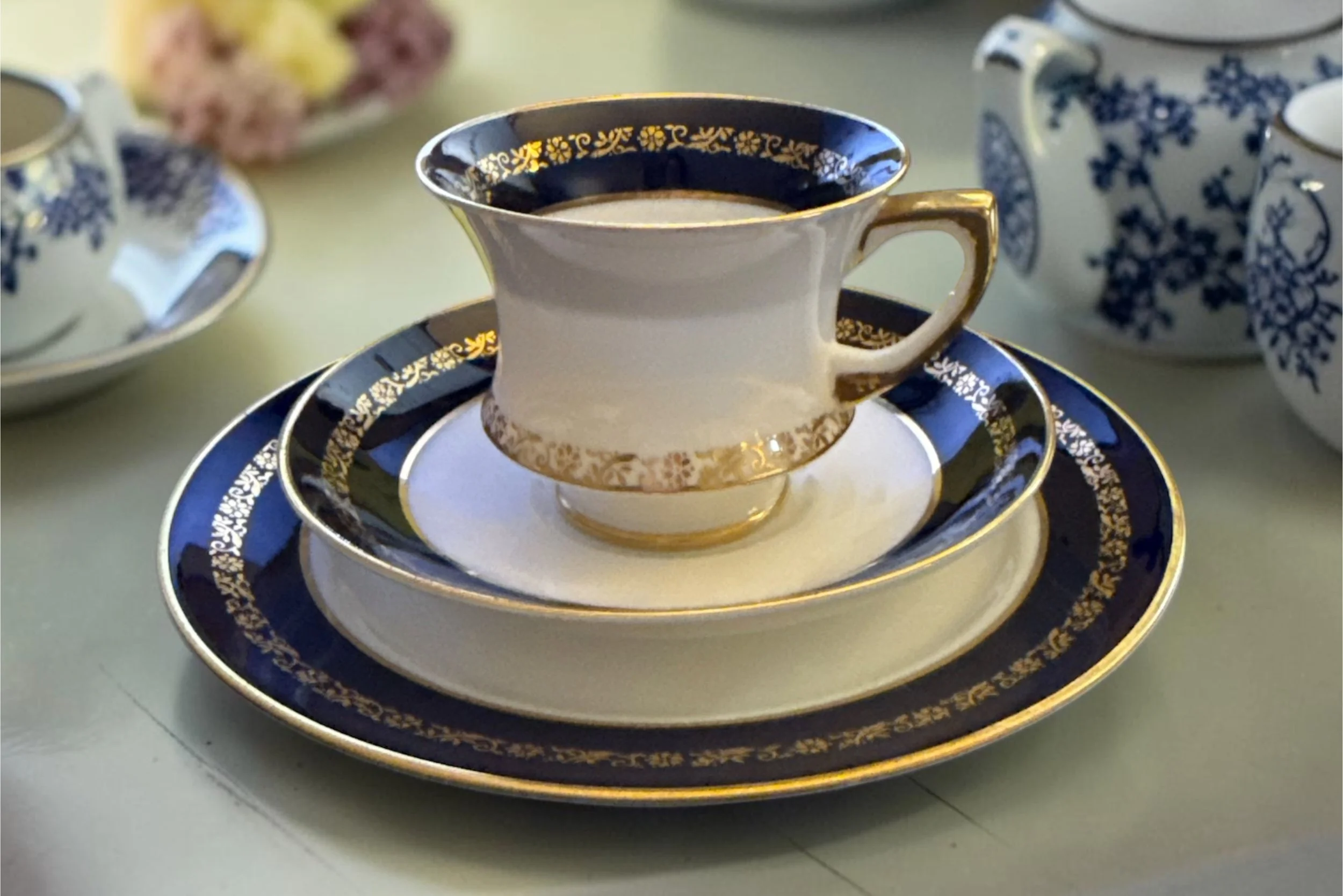 A porcelain tea cup and saucer with a navy blue and gold floral design, filled with a dark beverage, on a matching navy and gold patterned plate, with additional teapots and cups in the background.