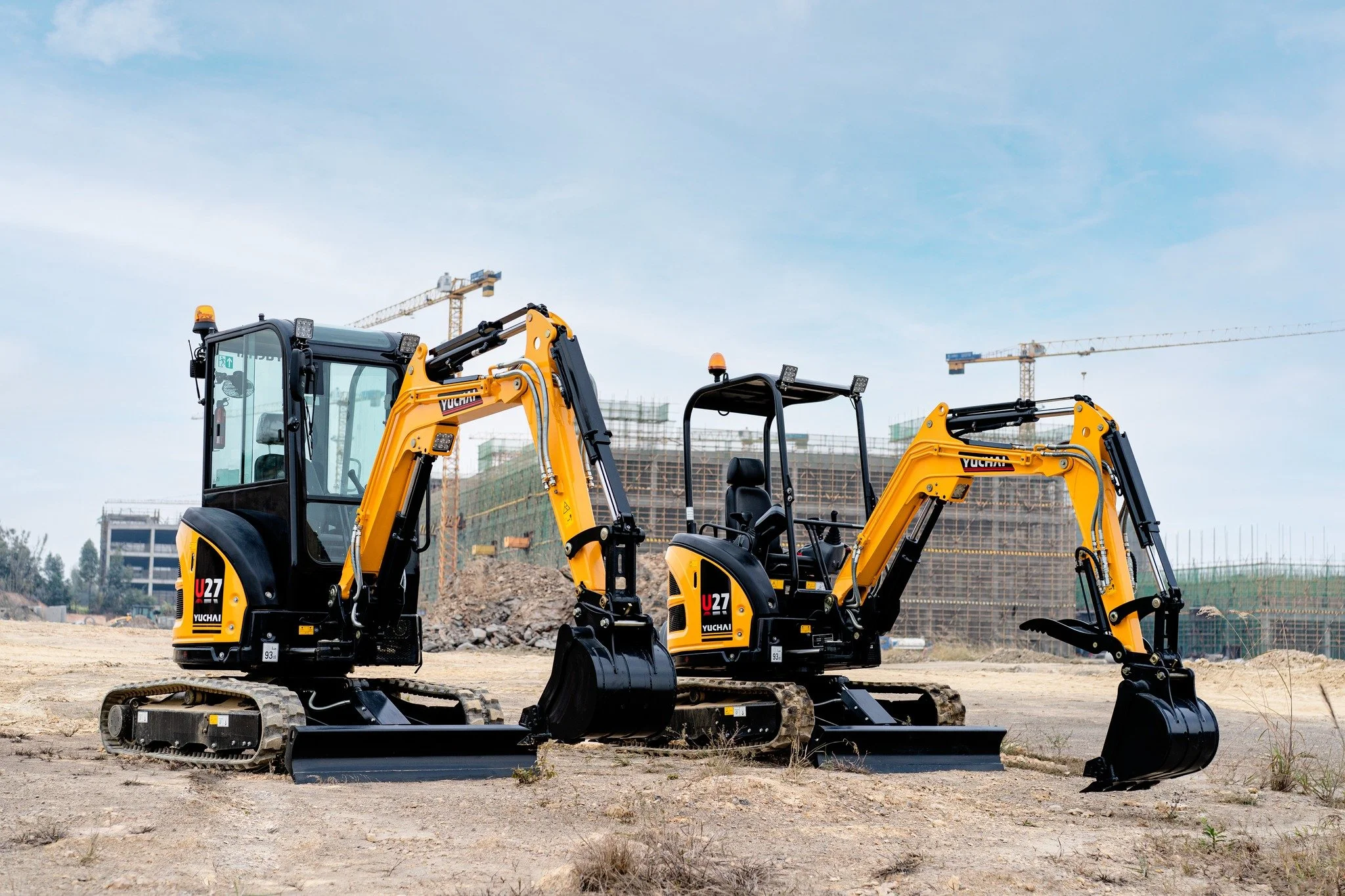 Two small yellow excavators with caterpillar tracks parked on a construction site with dirt and rocks, cranes and building structures visible in the background under a clear sky.
