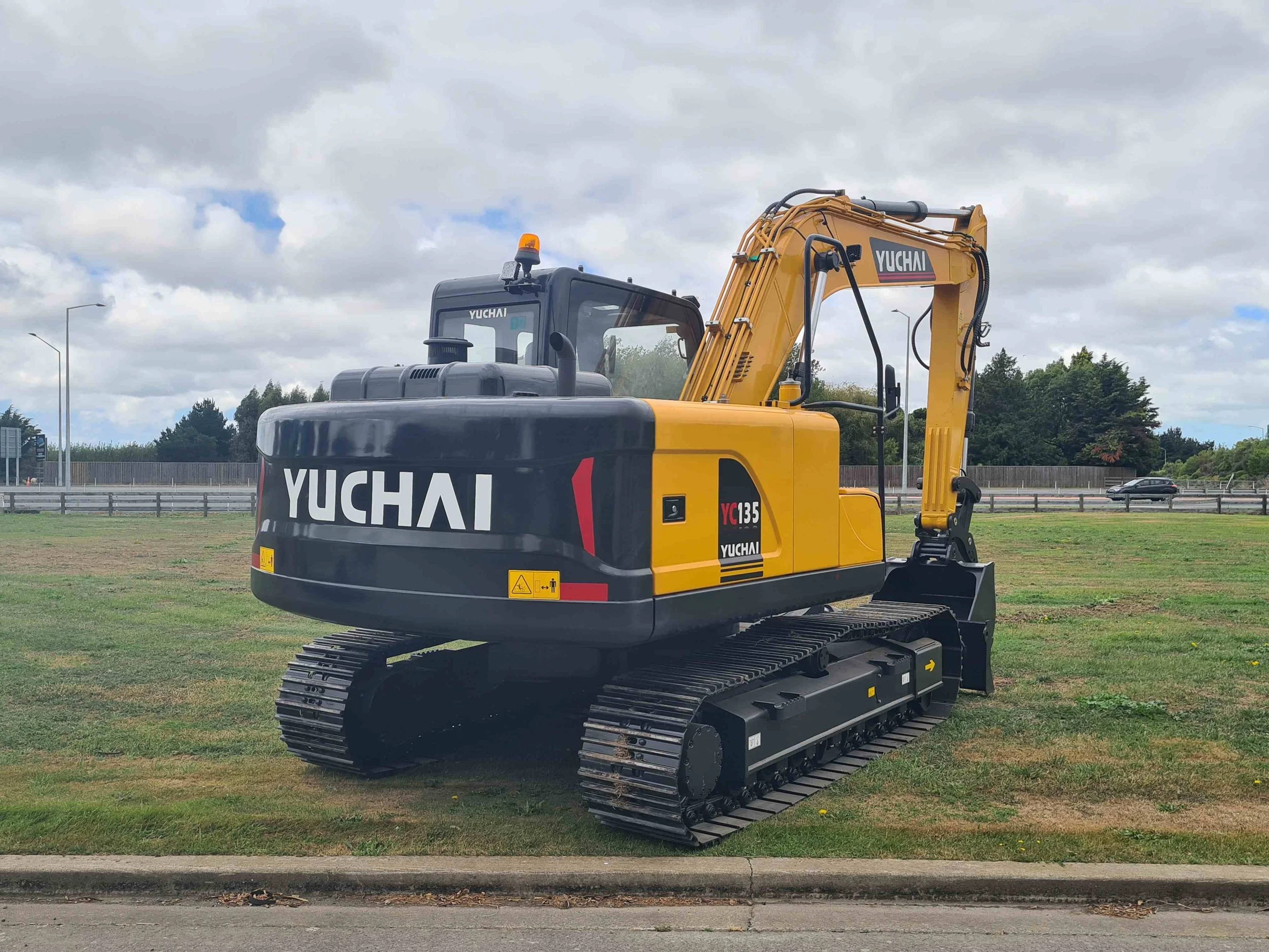 A yellow and black Yuchai excavator with rubber tracks on a grassy field under a cloudy sky.