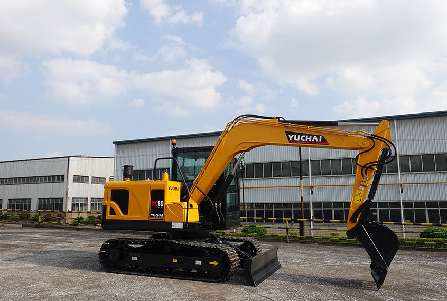 Yellow Yuchai excavator with a black blade and tracked wheels parked in front of industrial buildings.