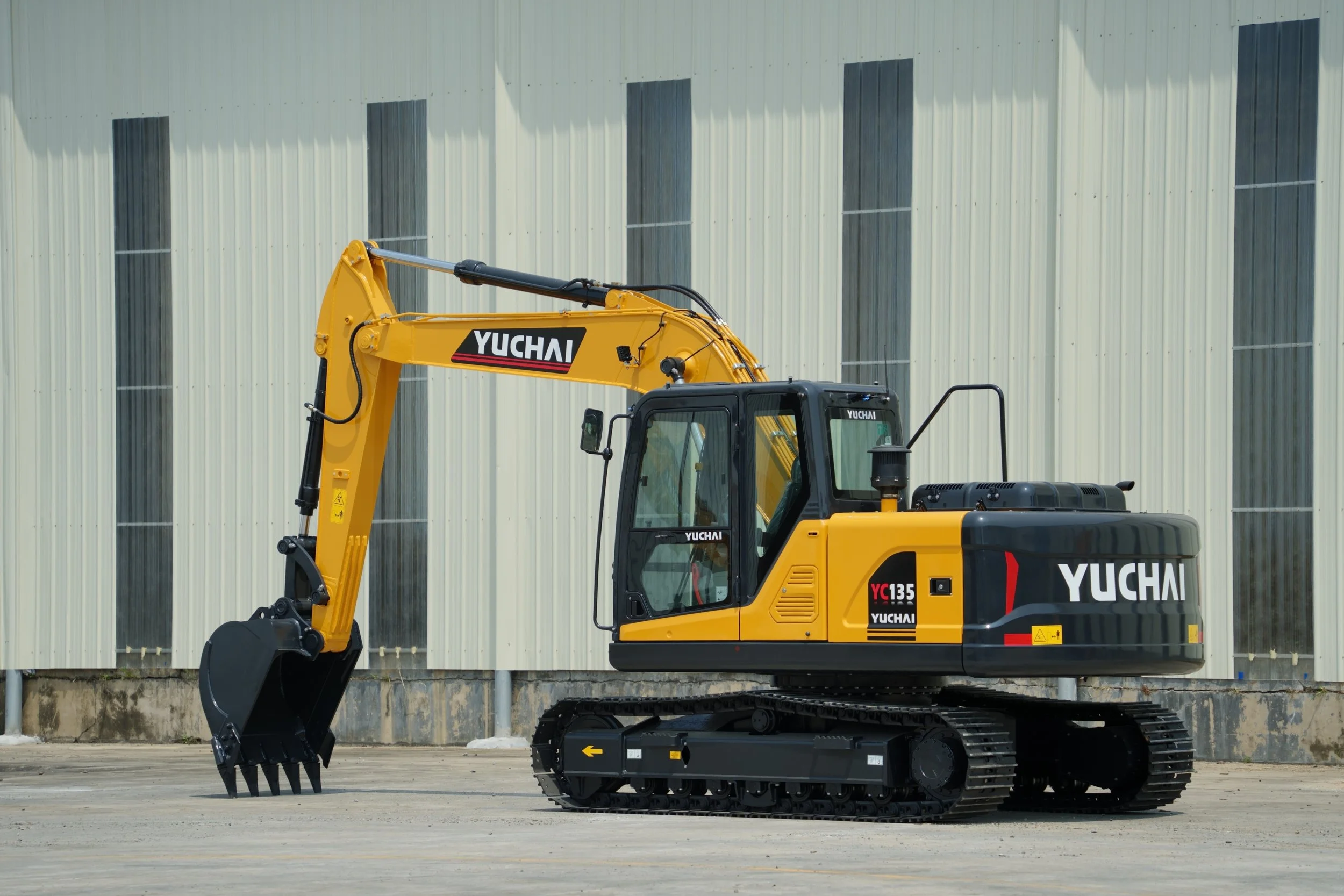 A yellow Yuchai excavator with black tracks parked on a concrete surface in front of a large corrugated metal building.
