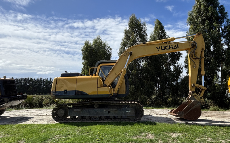 Yellow Yuchai excavator parked on a gravel surface with trees and a cloudy sky in the background.