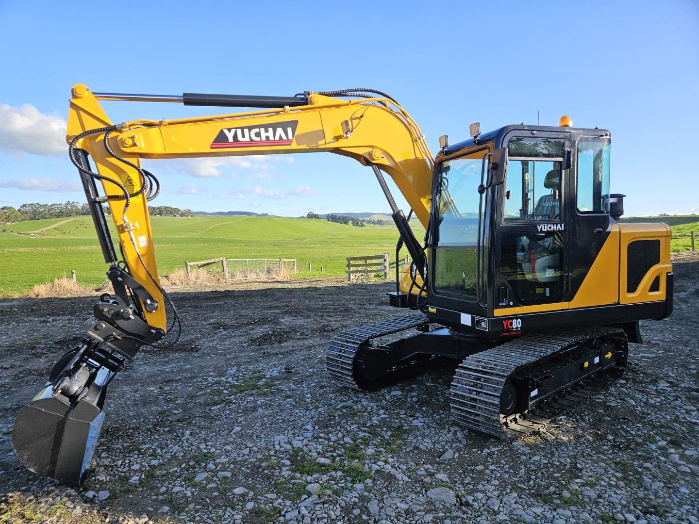 Yellow and black Yuchai excavator on rocky ground with green rolling hills and blue sky in the background.