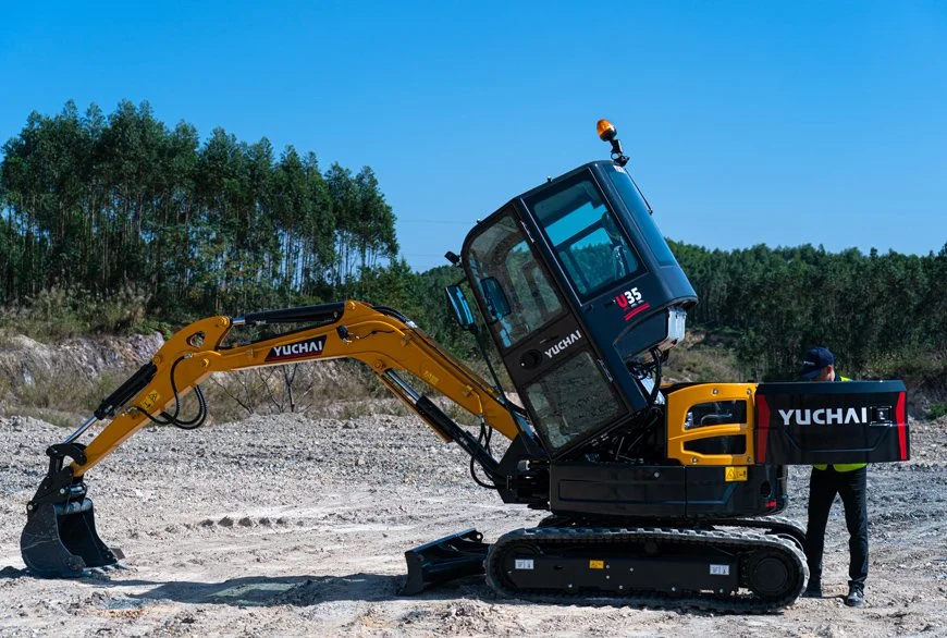A Yuchai mini excavator on a construction site with a person standing nearby, with trees in the background and a clear blue sky.