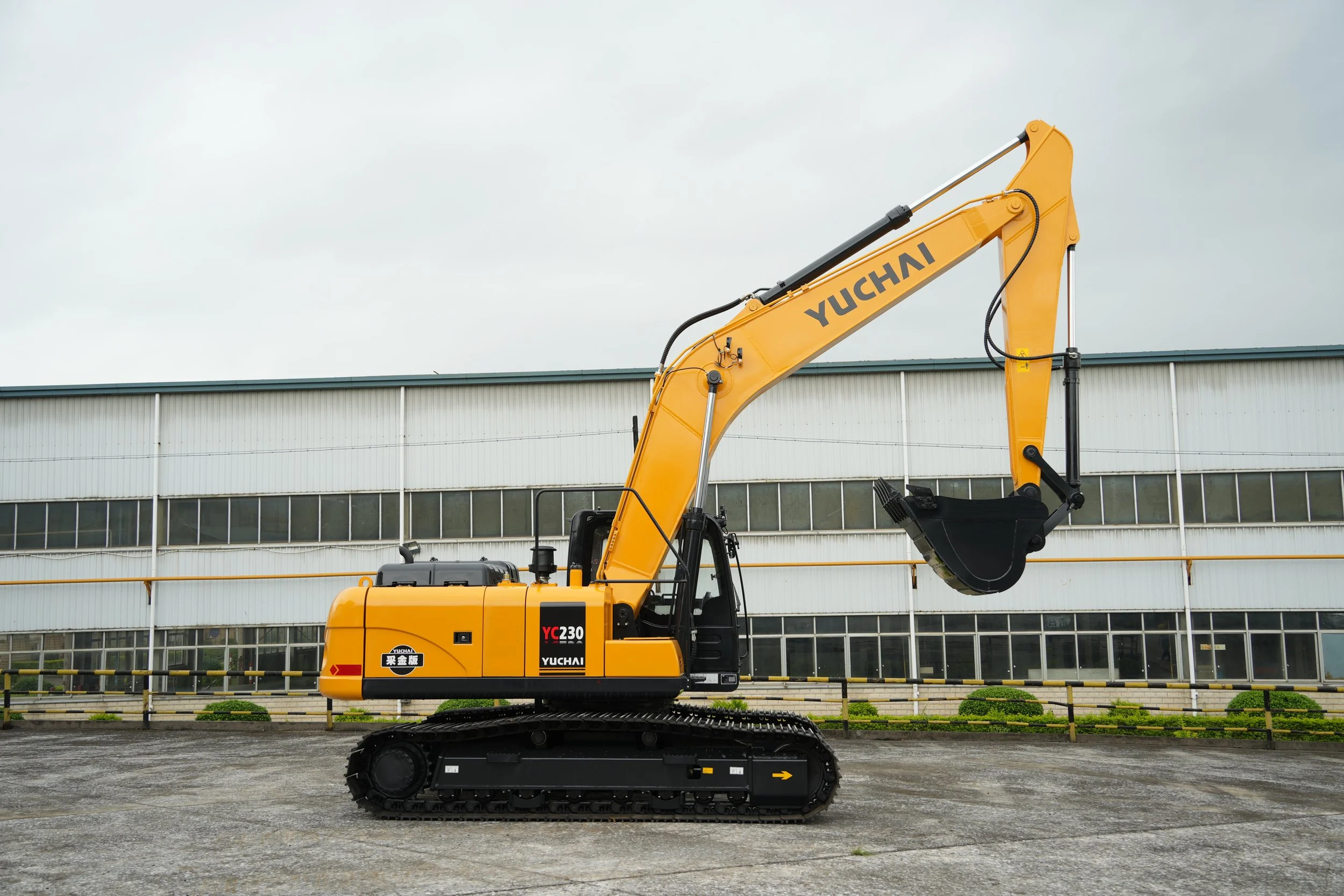 A yellow Yuchai YC230 excavator parked outdoors in front of a large industrial building with a gray roof and windows.