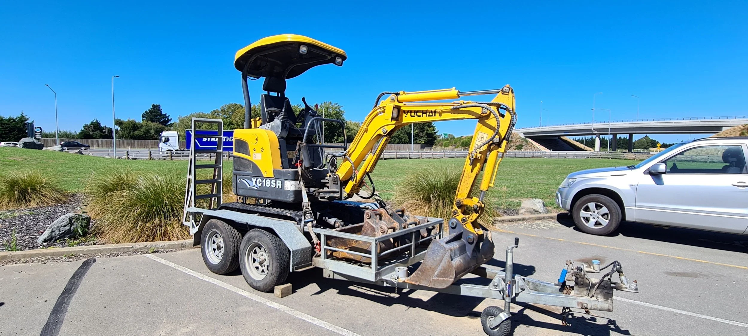 A yellow compact excavator parked in a parking lot next to a silver car, with a grassy area and a highway overpass in the background on a clear, sunny day.