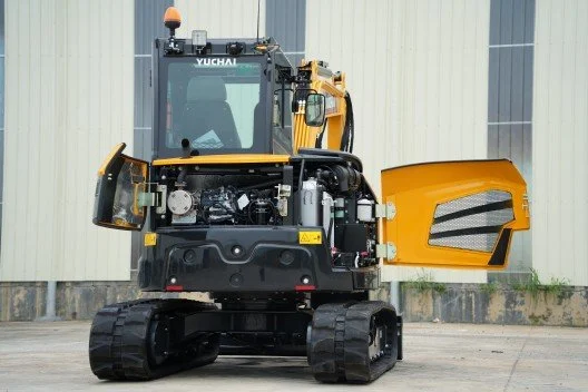 A yellow and black compact excavator with tracks, parked outdoors against a beige industrial building.