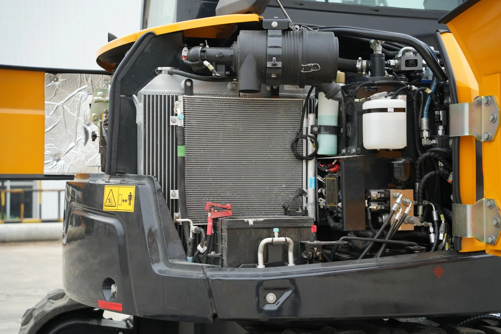 Close-up of an industrial machine's internal components, including a radiator, hoses, and various mechanical parts, with a yellow exterior and some warning labels.