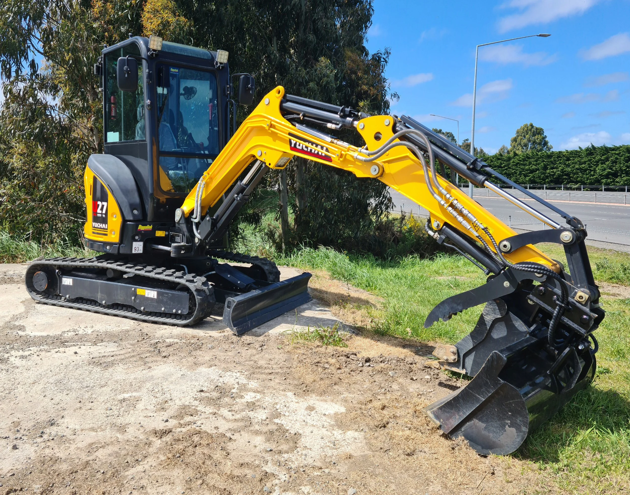 A yellow Yuchai excavator with black tracks parked on dirt next to green trees under a partly cloudy blue sky.