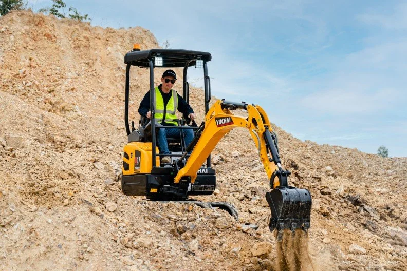 Person operating a small yellow excavator on a dirt mound at a construction site.