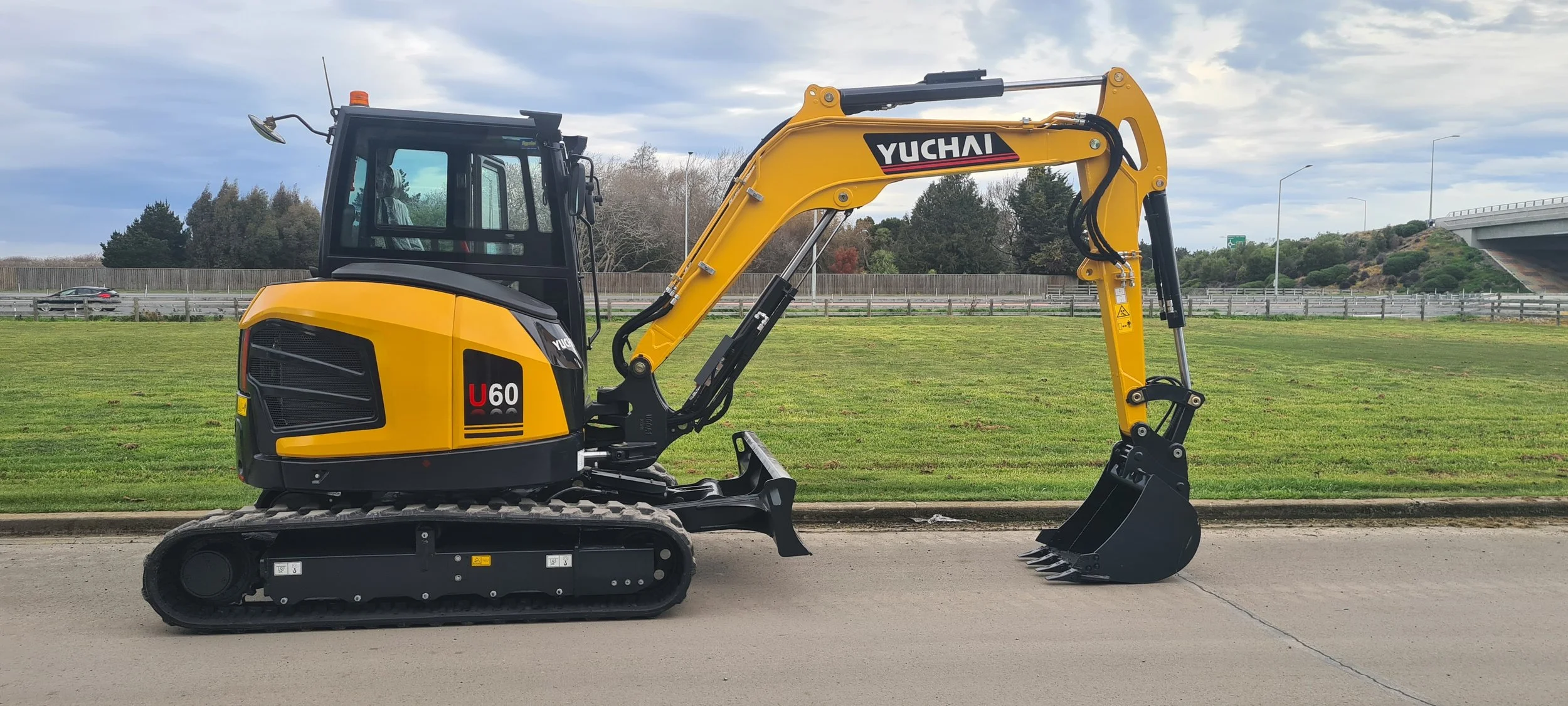 A yellow mini excavator with black tracks and arm, labeled "Yuchai U60" on a grassy area beside a paved road, with trees, fences, and a highway overpass in the background under a cloudy sky.