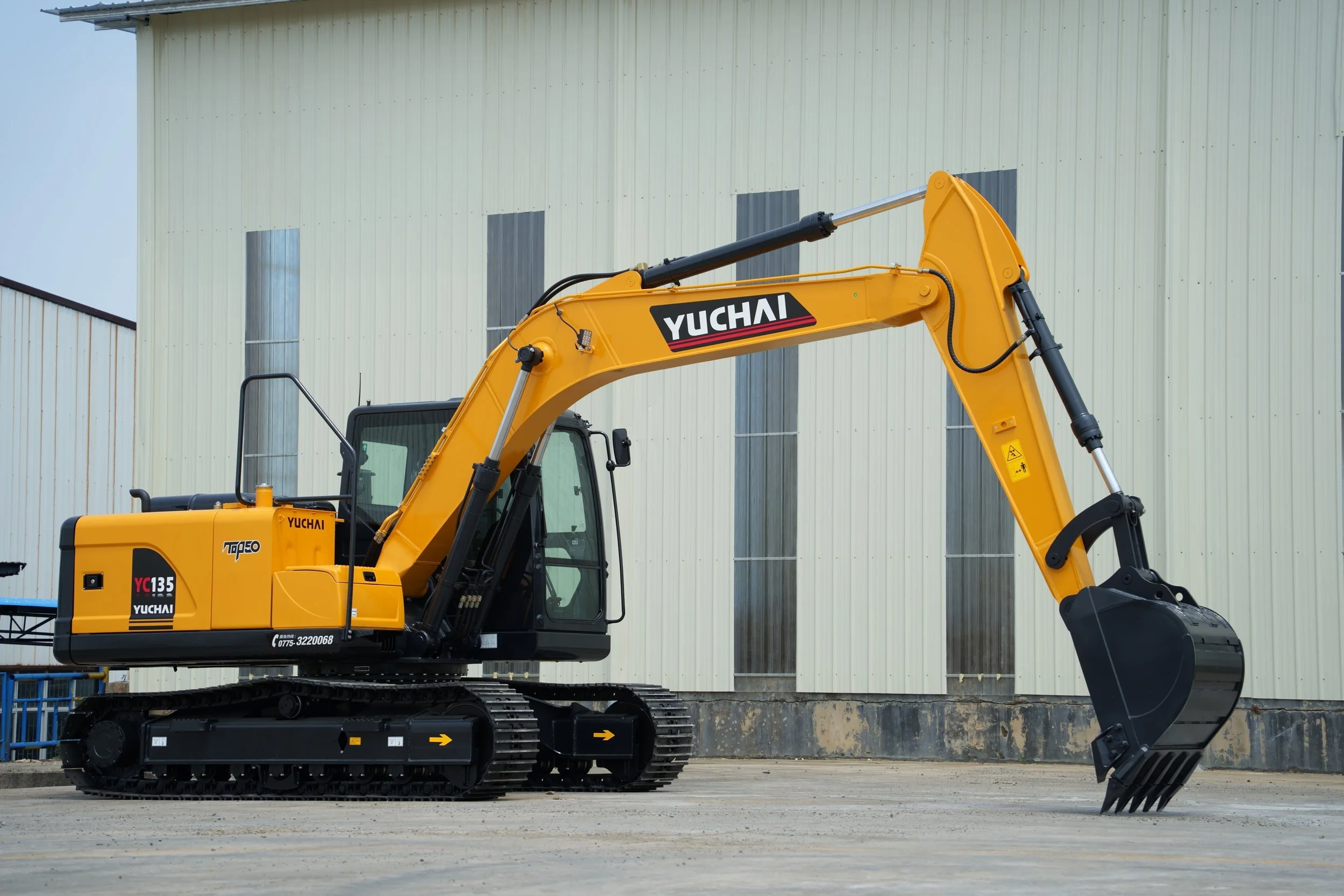 Yellow YUCHAI excavator parked on a concrete surface in front of a cream-colored industrial building with vertical windows.