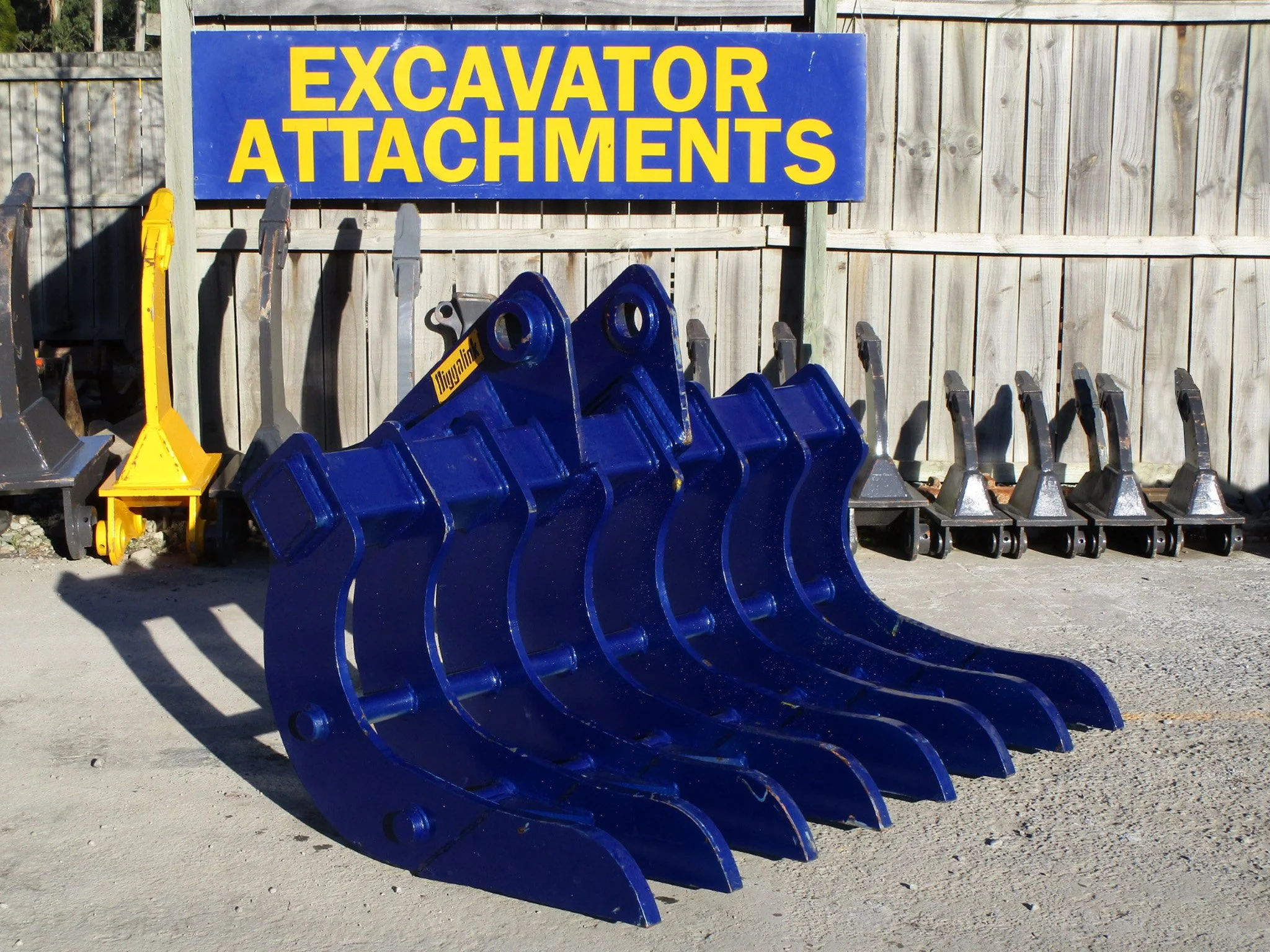 A row of blue excavator attachments, likely buckets, lined up outdoors in front of a wooden fence with a sign that reads "Excavator Attachments" in large yellow letters on a blue background.