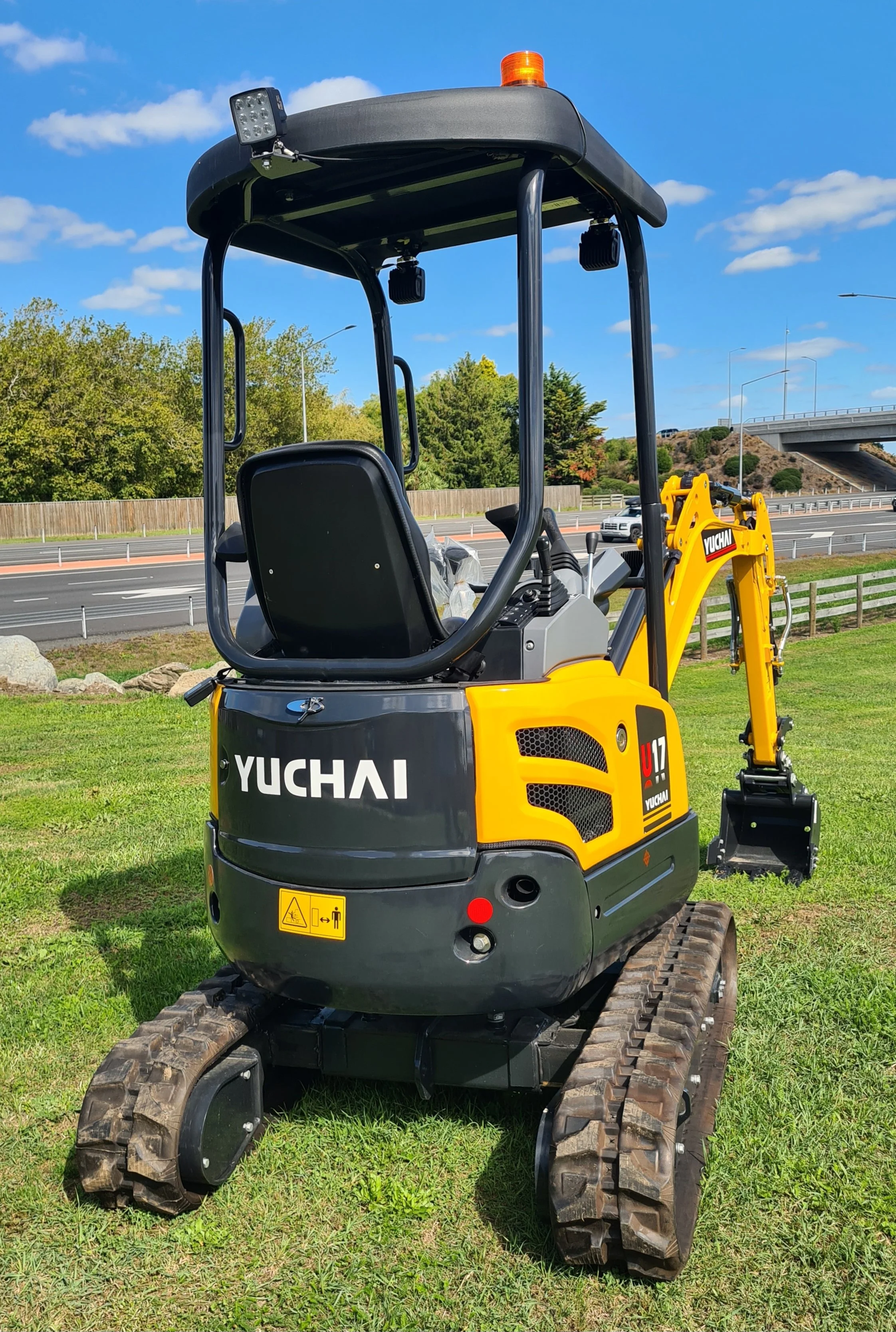 Compact Yuchai mini excavator with black and yellow body, rubber tracks, and a small bucket attachment, situated on a grassy area near a road with green trees in the background.