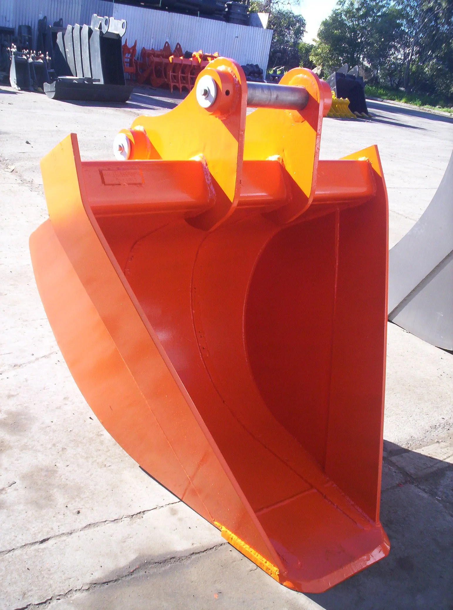 Close-up of a large orange snowplow blade with mounted metal pole, with various equipment stored in the background outdoors.