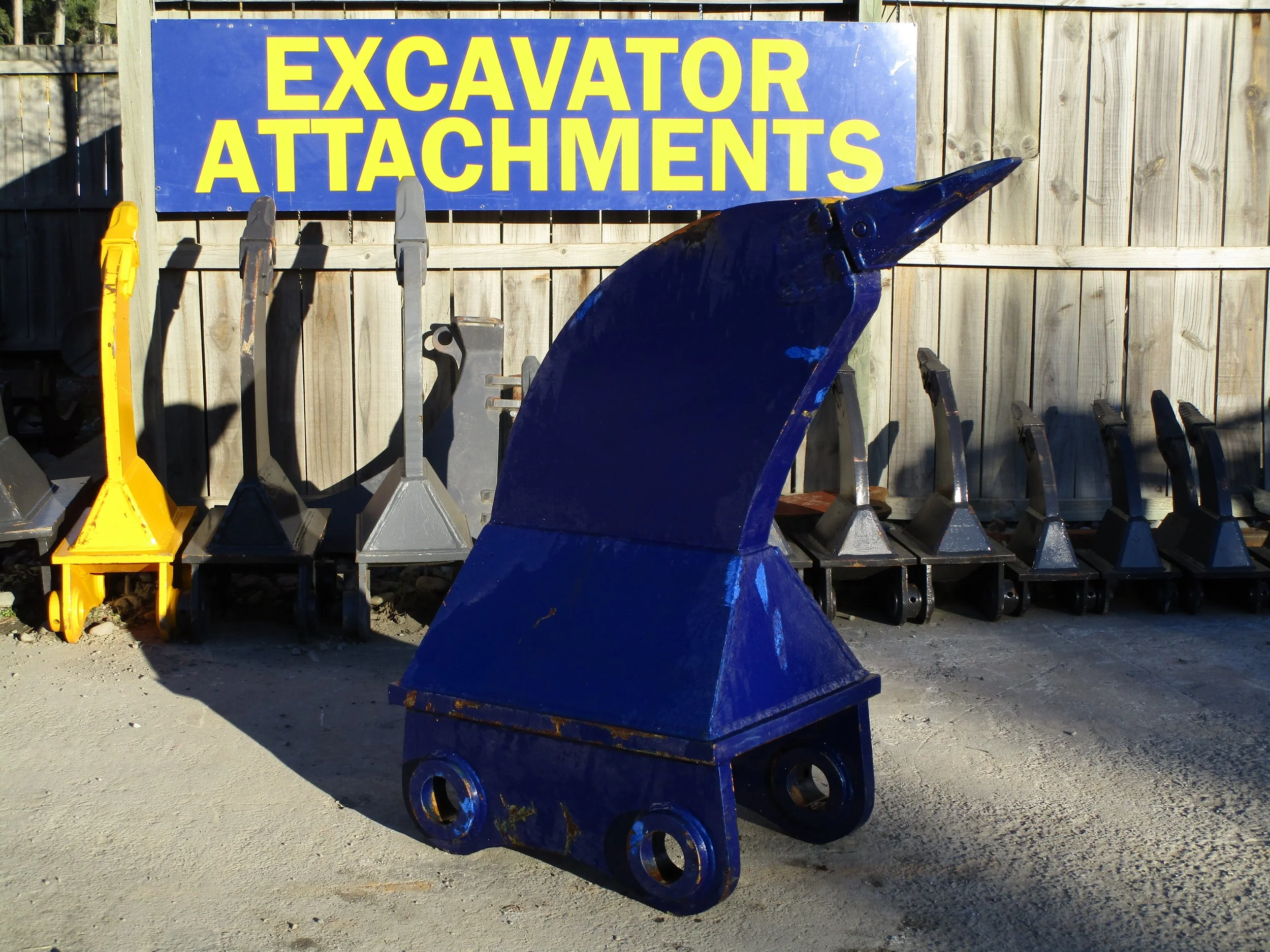A blue excavator attachment sitting on the ground with a yellow sign in the background that reads 'EXCAVATOR ATTACHMENTS' and a wooden fence.