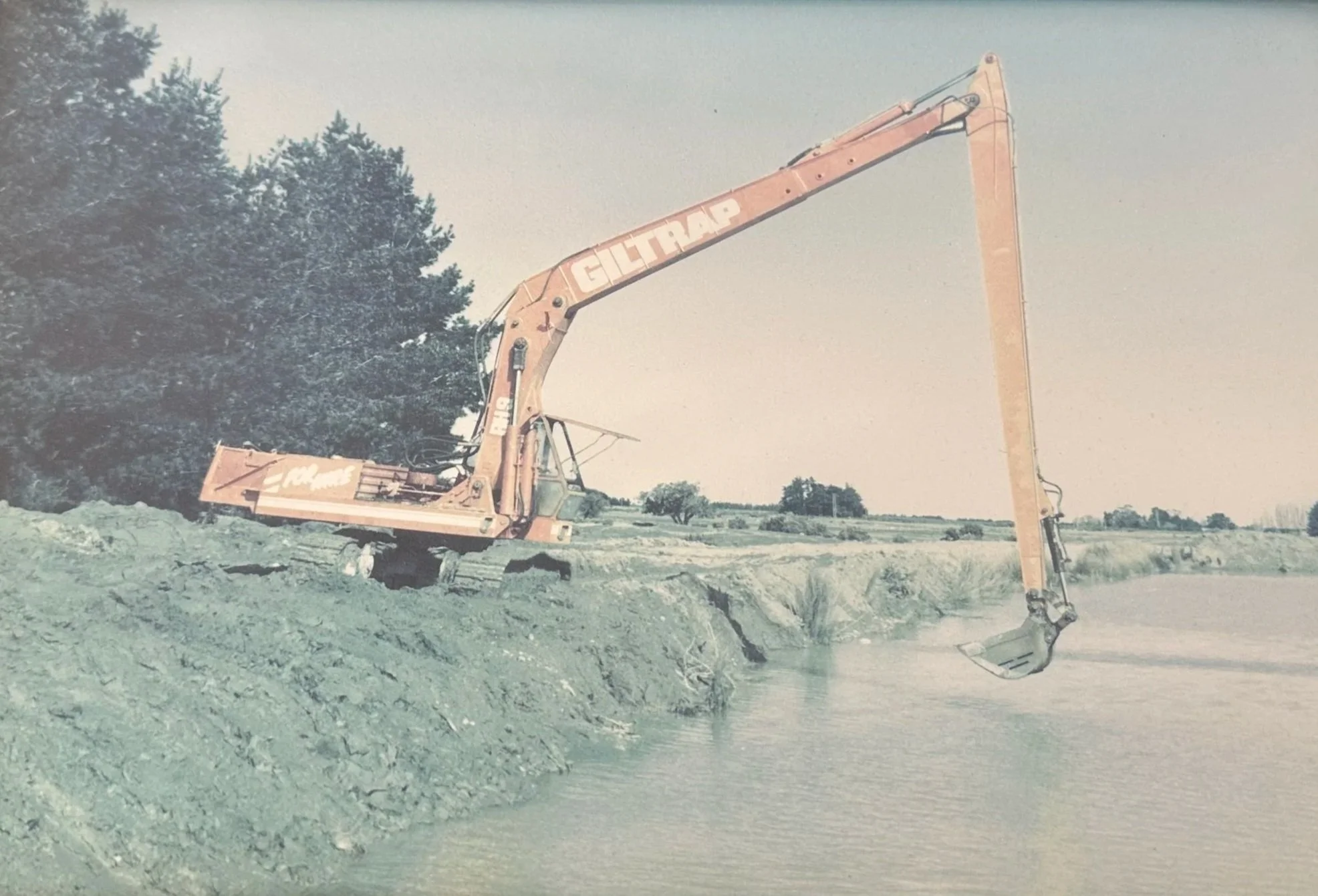 An orange excavator with the brand name 'GILTTRAP' on its arm, parked on a construction site with dirt and rocks, near some trees in the background.
