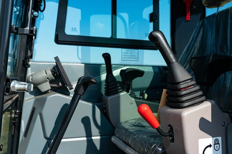 Interior of a construction vehicle cabin with two control levers, one with a red handle, and a screen on the dashboard.