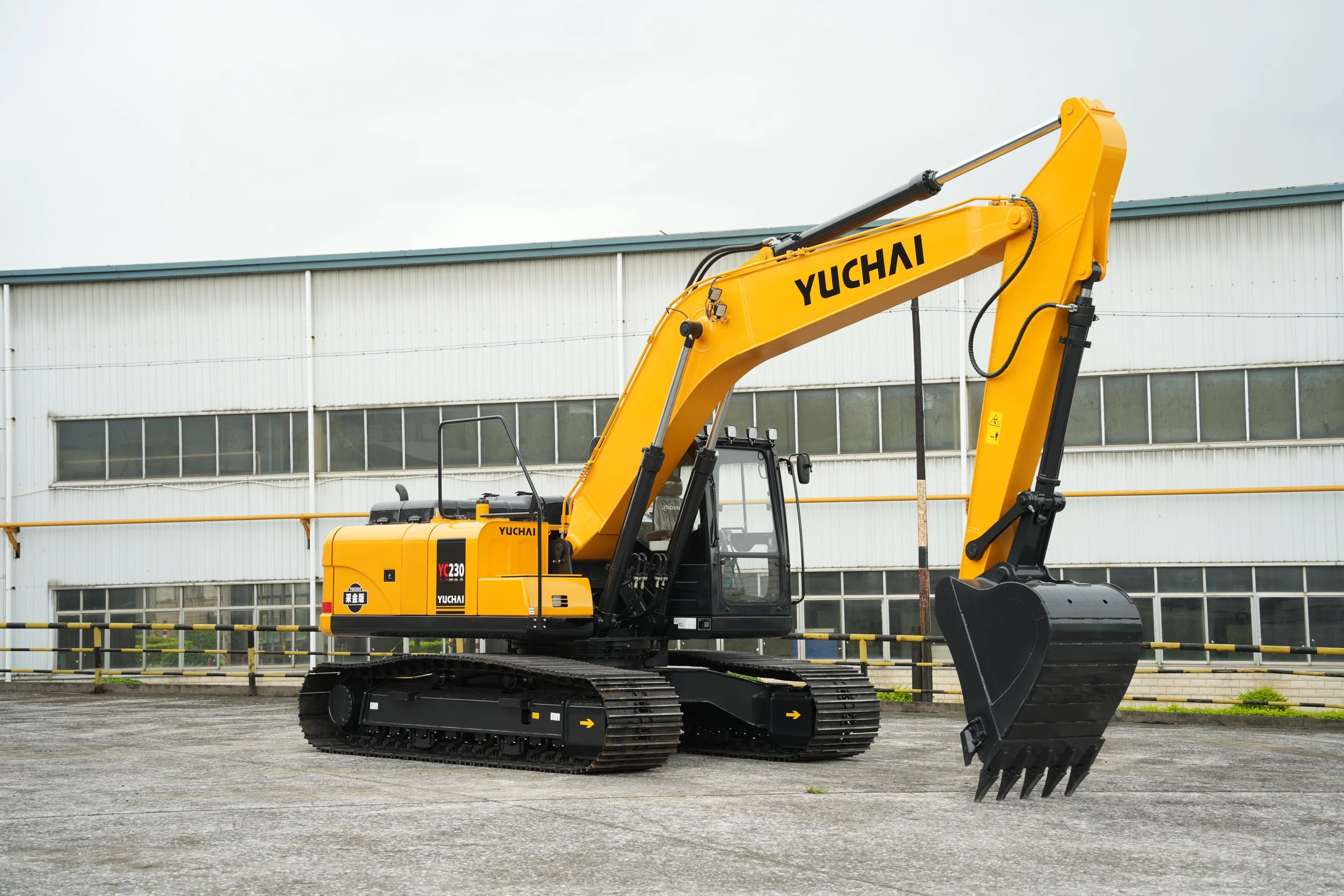 A yellow Yuchai excavator with black tracks parked outdoors near a modern industrial building with large windows.