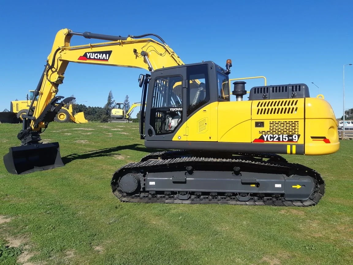Yellow and black excavator with the brand name YUCHAI on the arm and model YC215-9, positioned on a grassy field under a clear blue sky.