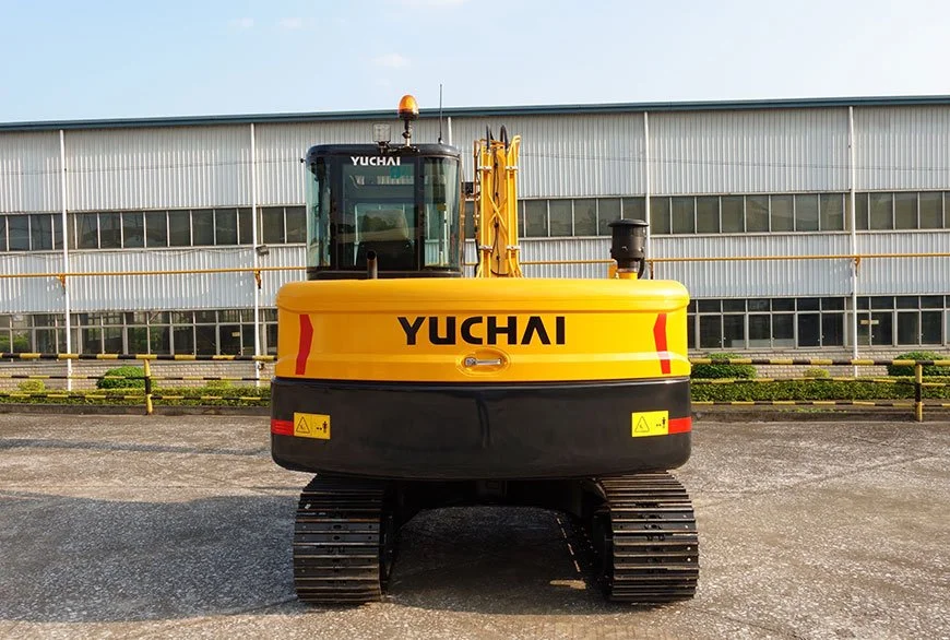 Yuchai compact yellow excavator with black undercarriage, parked in front of a large industrial building with multiple windows.