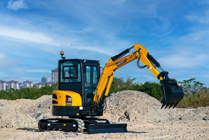 Small yellow and black excavator on dirt with construction site and city skyline in background under blue sky.