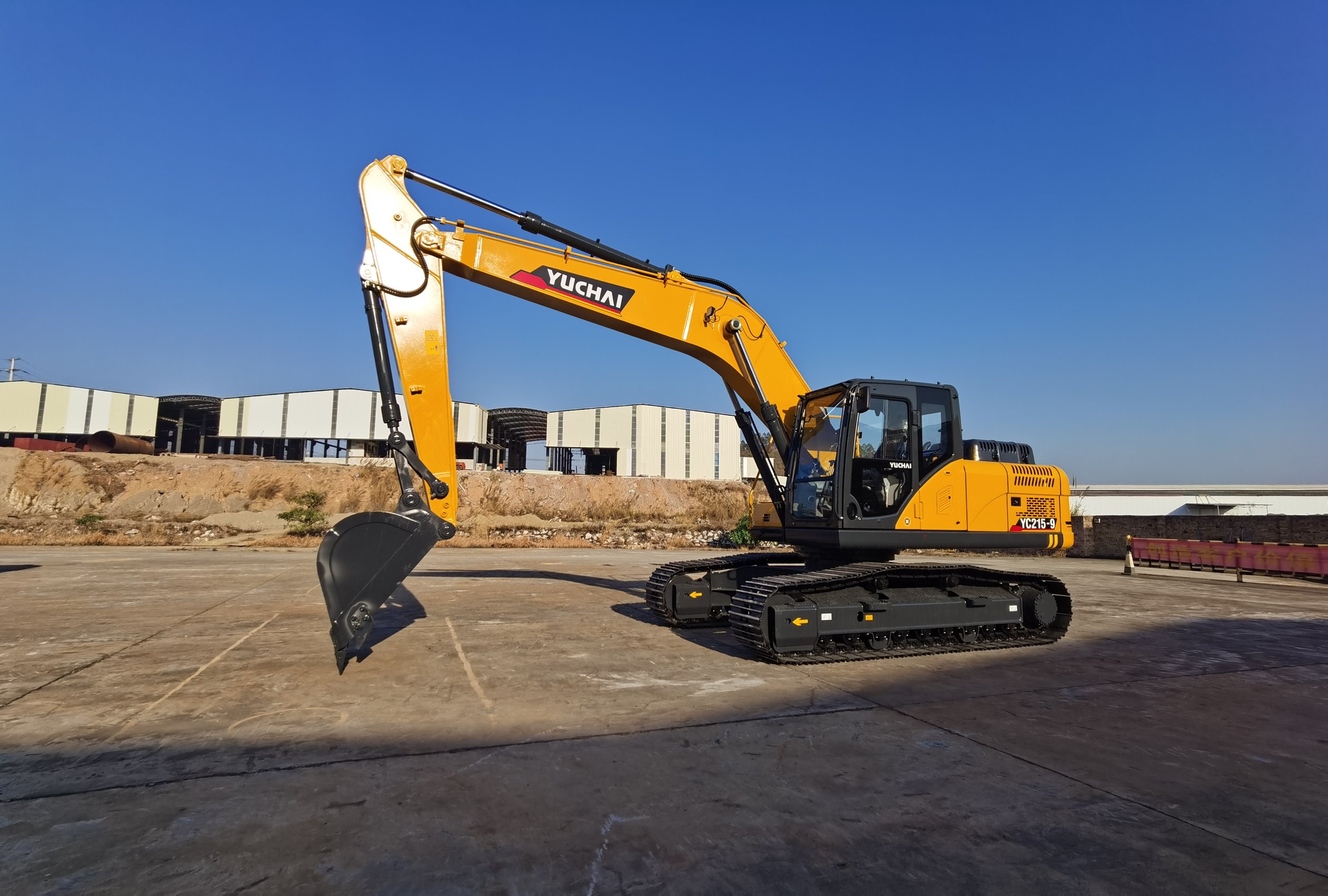 Yellow excavator on a construction site against a clear blue sky, with industrial buildings in the background.