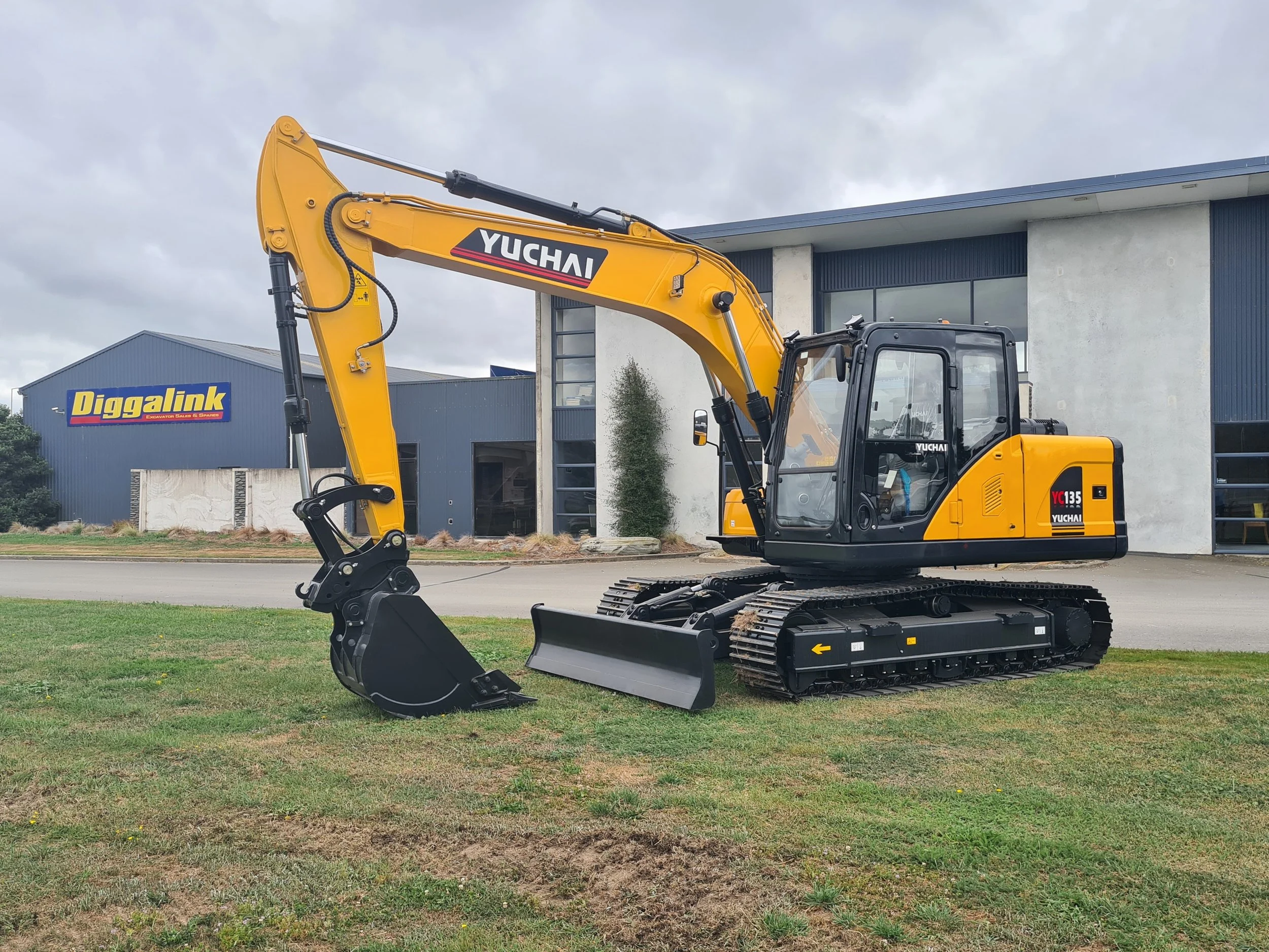 A yellow Yuchai excavator parked on a grassy area in front of buildings with signs, including one that says 'Diggalink'. The sky is cloudy.
