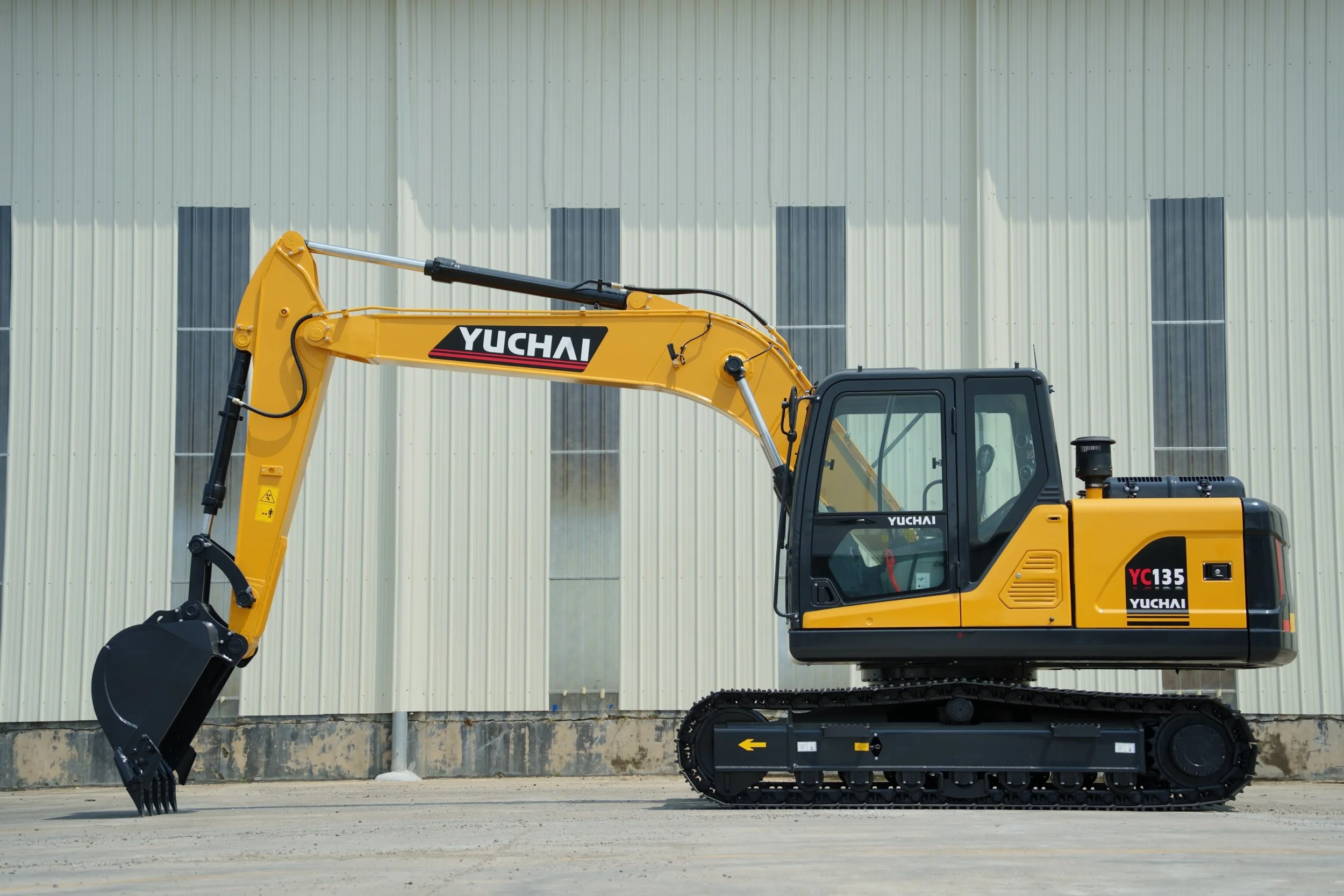 Yellow Yuchai excavator parked on a construction site with a large industrial building in the background.