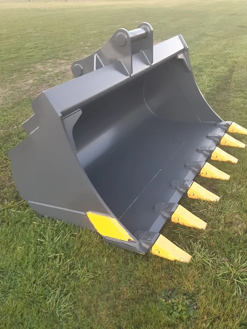 Close-up of a large gray excavator bucket with yellow teeth, resting on green grass.