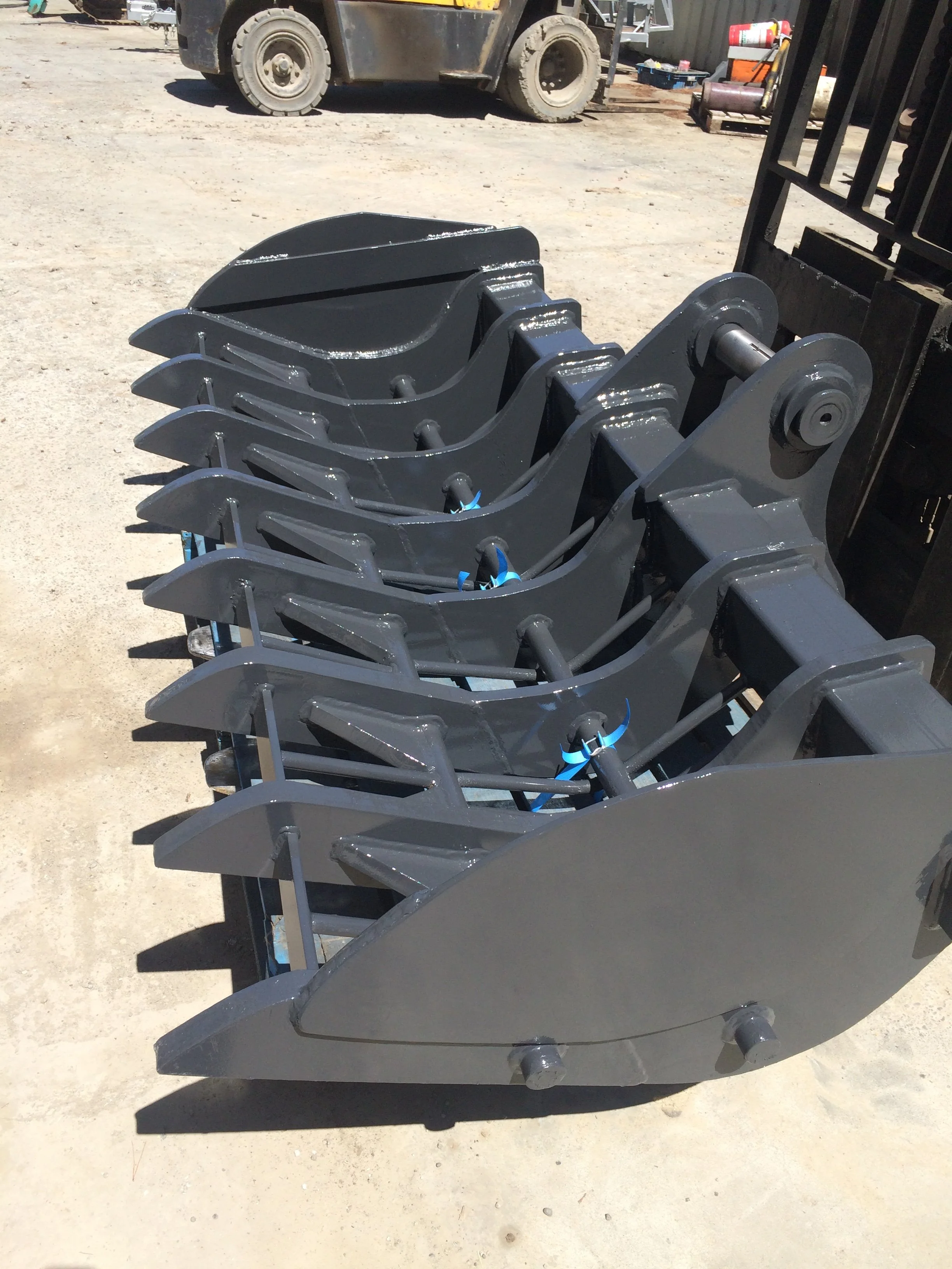 Close-up of a mechanical attachment or bucket for heavy machinery, painted black, with a background showing a dirt ground, a forklift, and various tools and supplies.