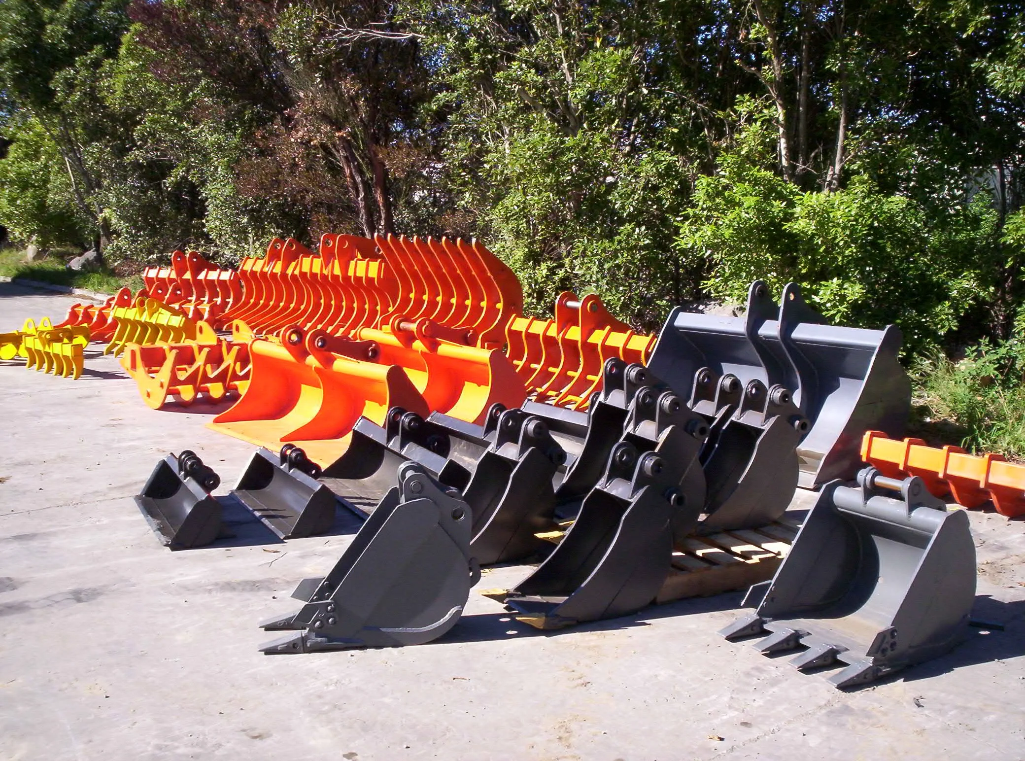 A line of construction or excavator buckets arranged outdoors with green trees and blue sky in the background.