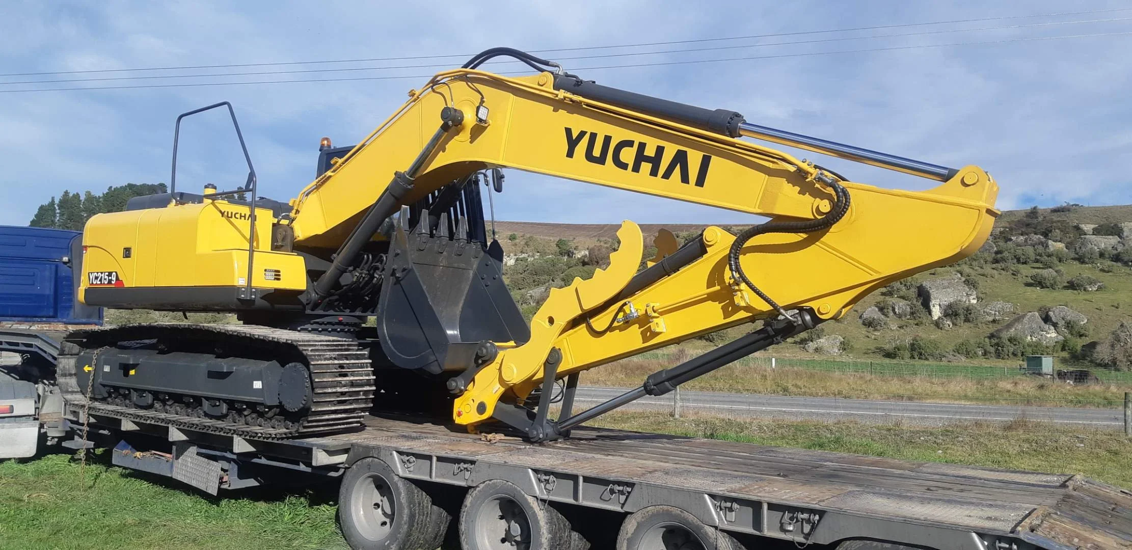 A yellow Yuchai excavator on a flatbed truck in a rural area with hills and trees in the background.