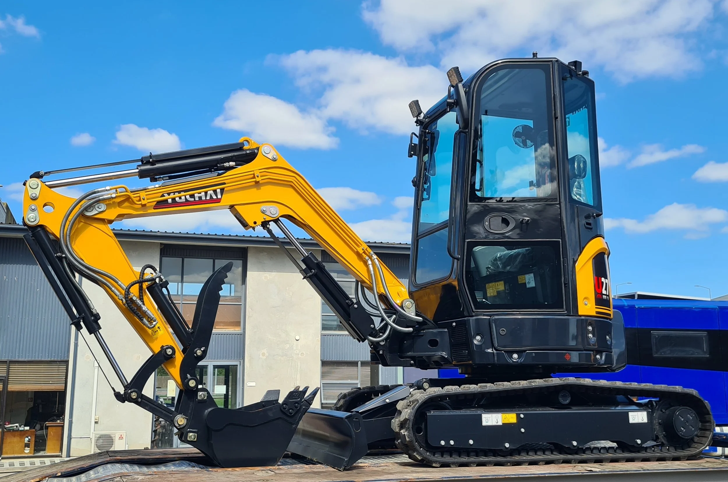 A yellow excavator with a black cab and tracks, parked outdoors under a blue sky with white clouds, in front of a modern building.