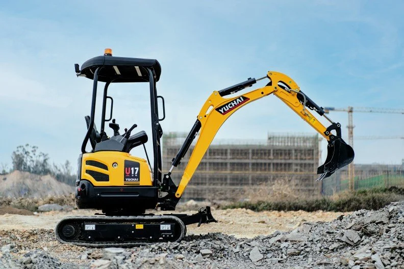 Yellow compact excavator parked on a construction site with a dirt mound in the foreground and construction in the background.