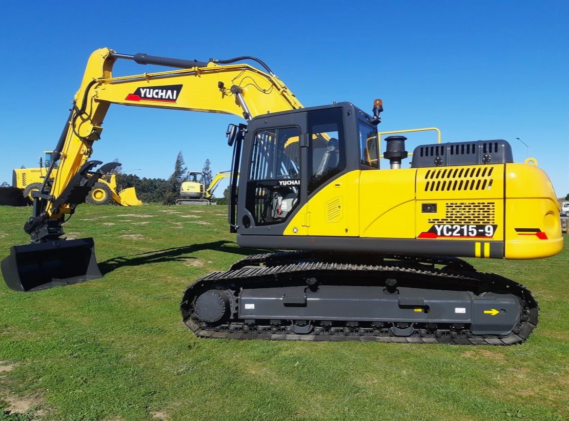 Yellow and black Yuchai excavator with tracked undercarriage on green grass field under clear blue sky, with other construction equipment in background.