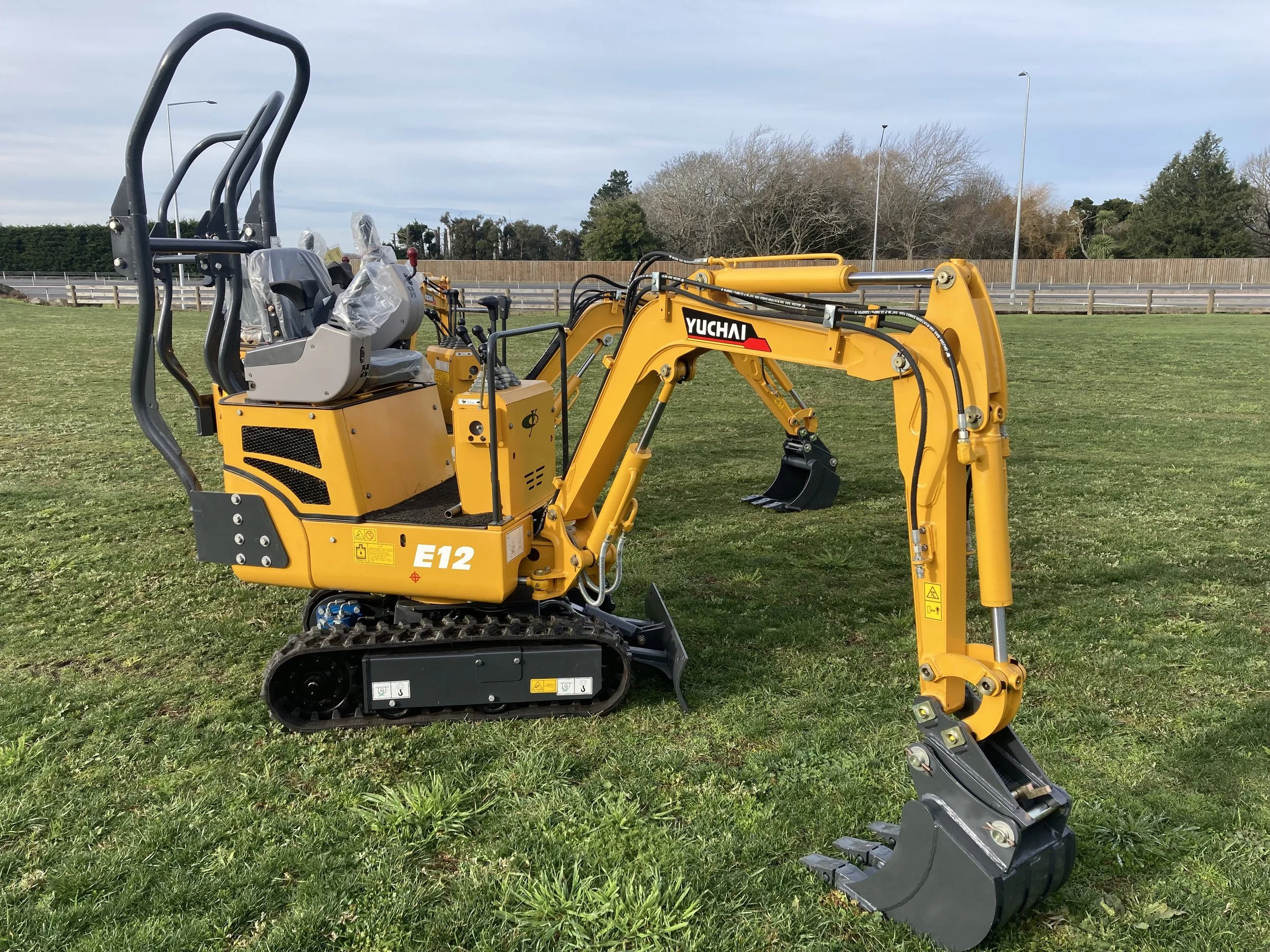 A yellow mini excavator with black rubber tracks, a small bucket, and a protective seat, standing on a grassy field near a fence and road, under a partly cloudy sky.