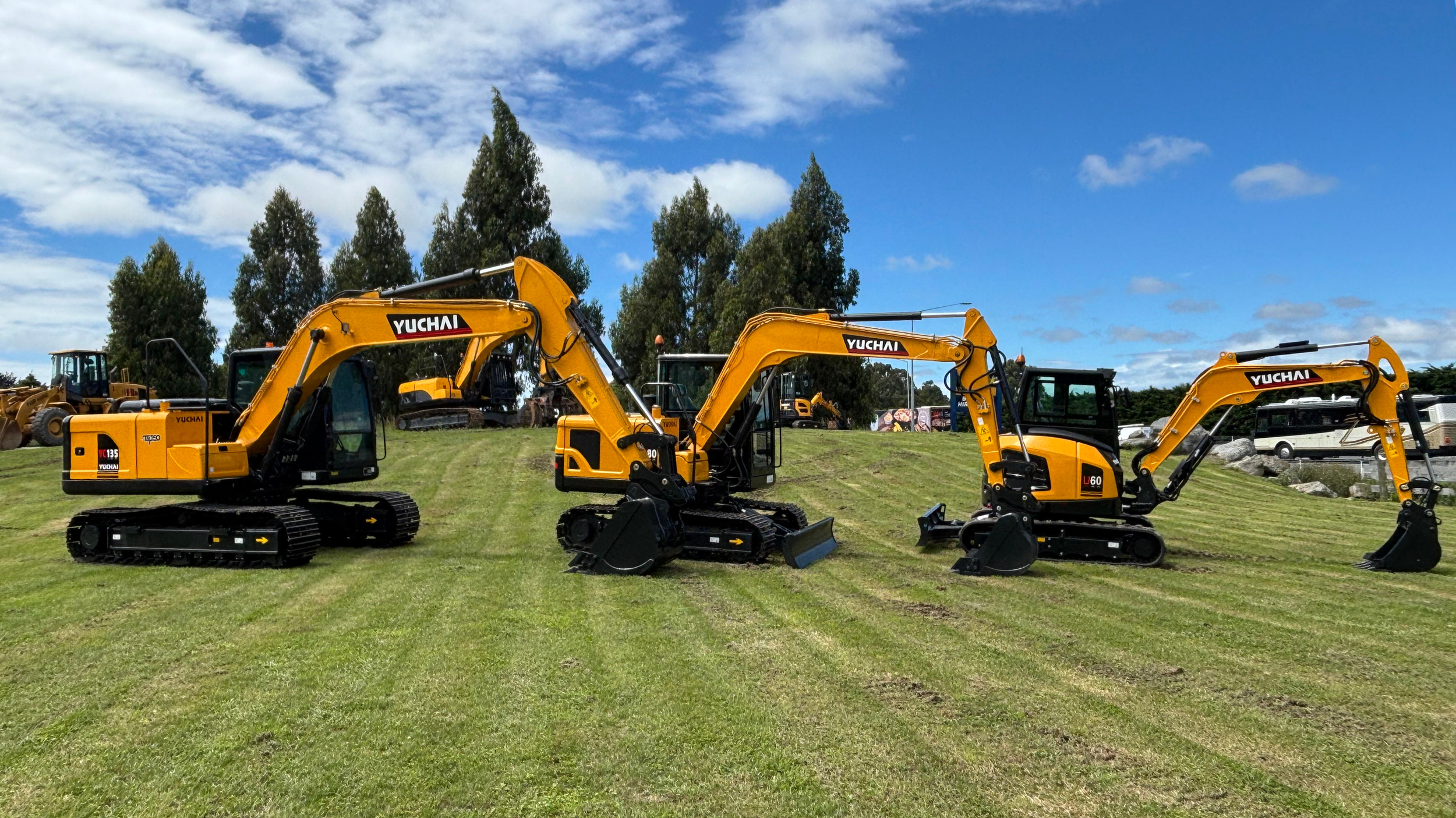 Three small yellow excavators with black tracks, labeled 'YUCHAI,' lined up on a well-maintained grassy field with a row of tall trees and a partly cloudy blue sky in the background.