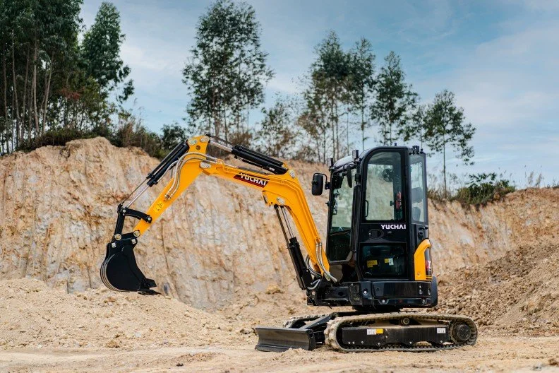 Small yellow and black excavator on a dirt construction site, with a dirt embankment and trees in the background.