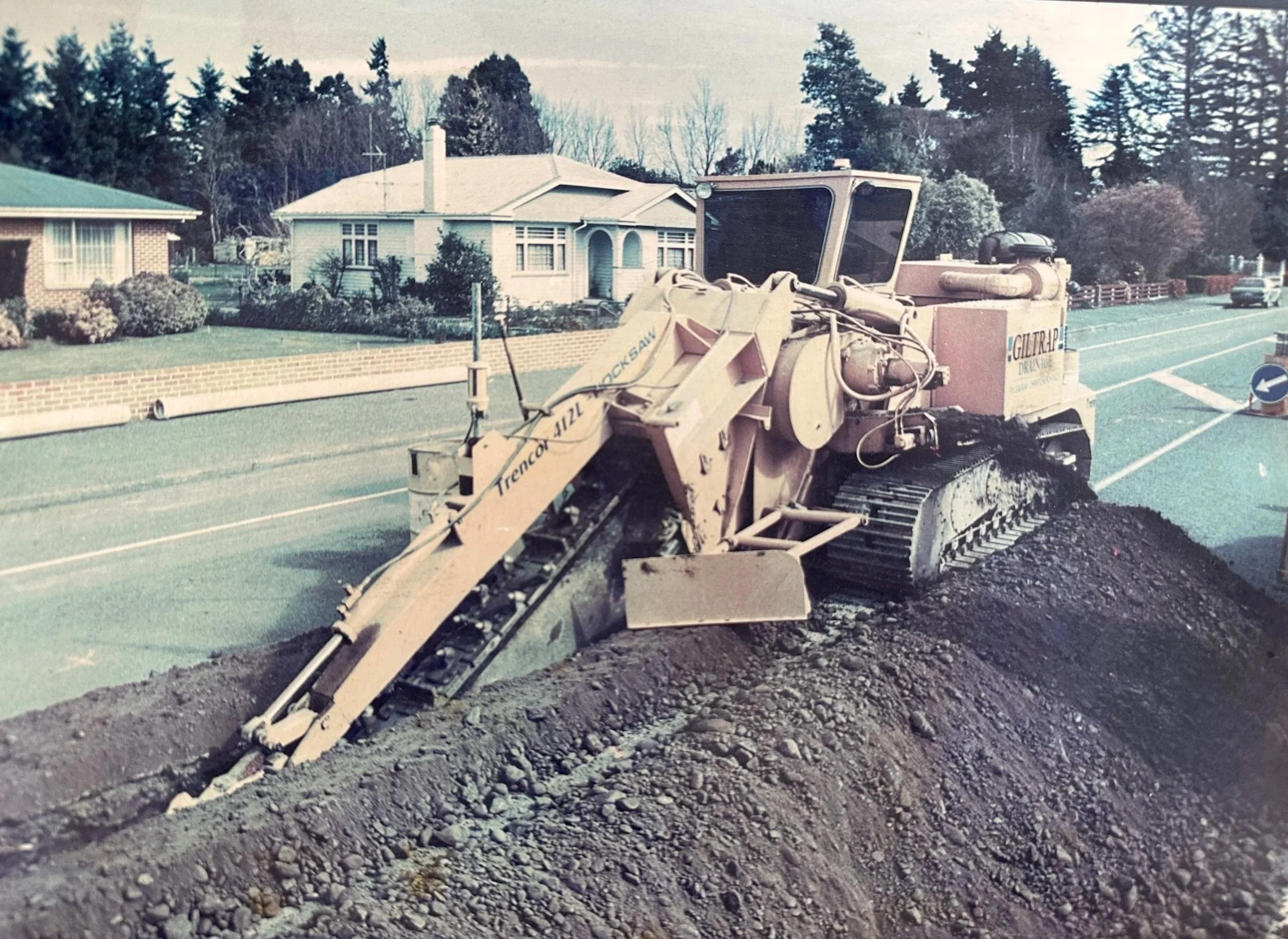 A road construction site with a large asphalt paver machine working on the road, in a suburban neighborhood with houses, trees, and parked cars in the background.