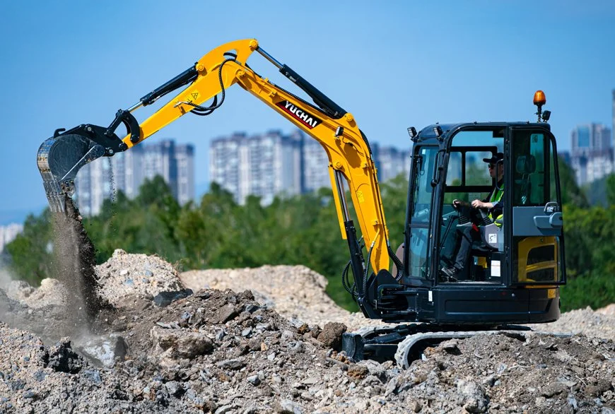 A yellow and black excavator with the brand logo 'Yuchai' is moving dirt on a construction site, with urban buildings in the background.