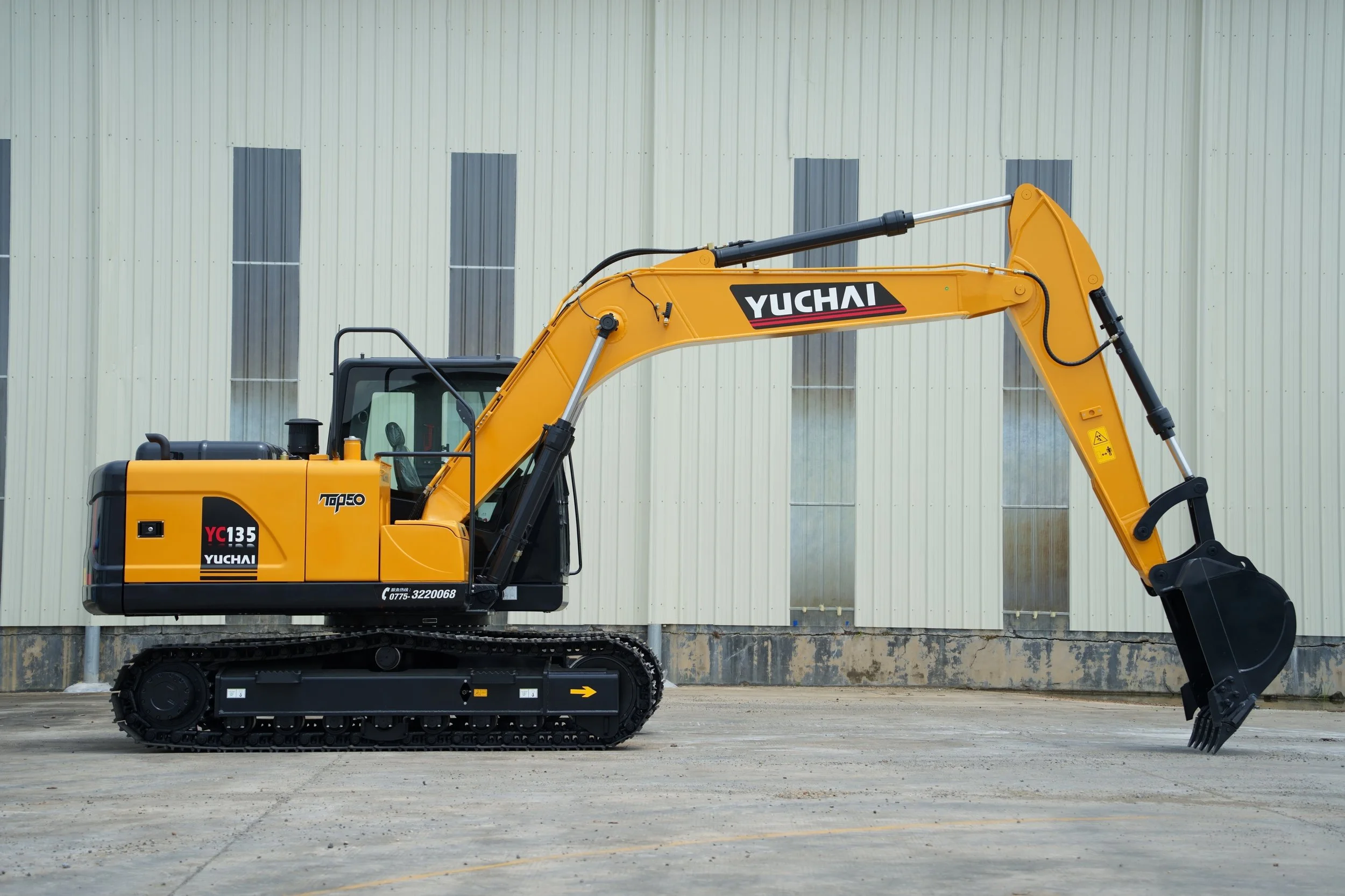 Yellow Yuchai excavator parked on a concrete surface in front of a beige industrial building with vertical metal siding.