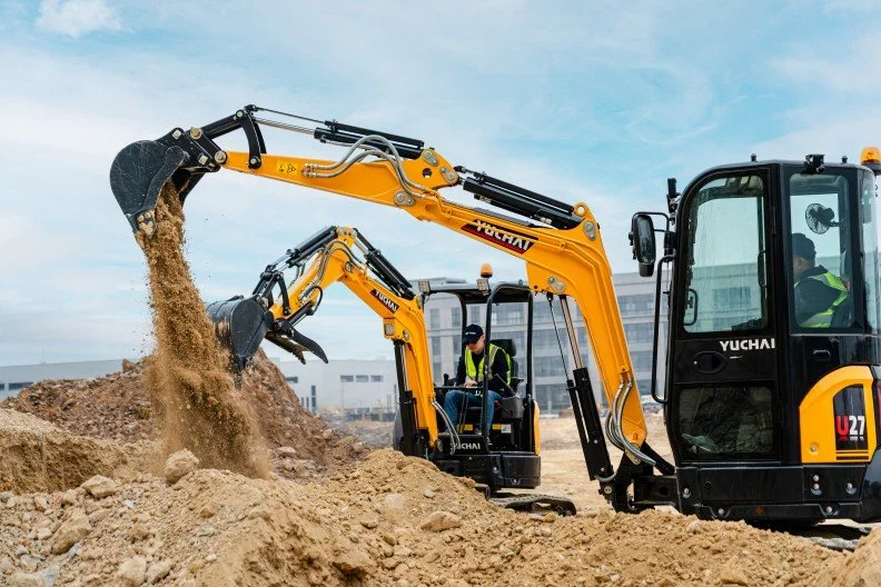 Construction site with two yellow excavators digging into a dirt mound, with workers operating the machinery and wearing safety vests and helmets.