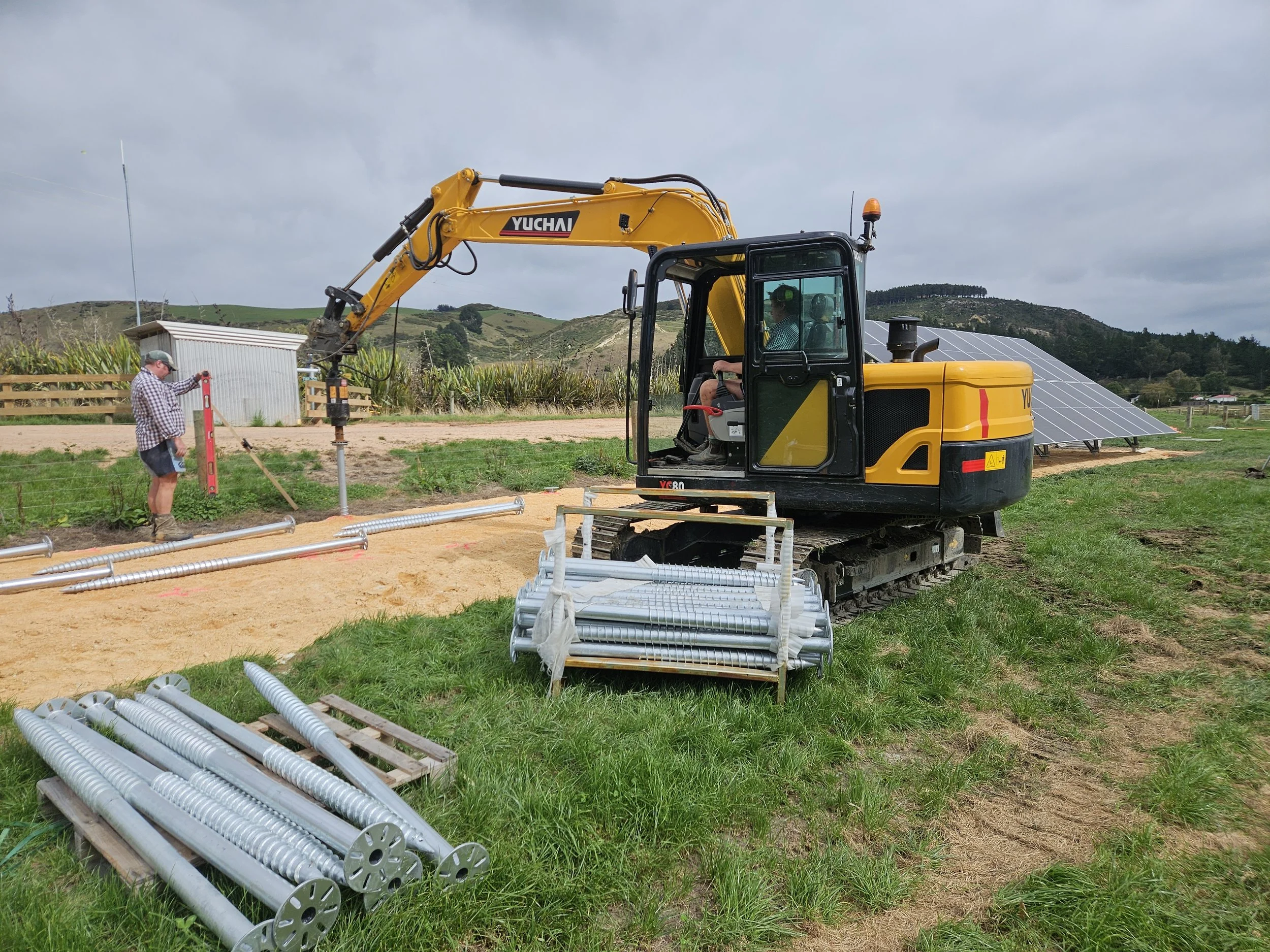 A small yellow excavator digging in a grassy field with mountains in the background. A person in a checkered shirt and cap operates the excavator, while another person in sunglasses and a cap observes nearby. Solar panels are visible on a hillside in