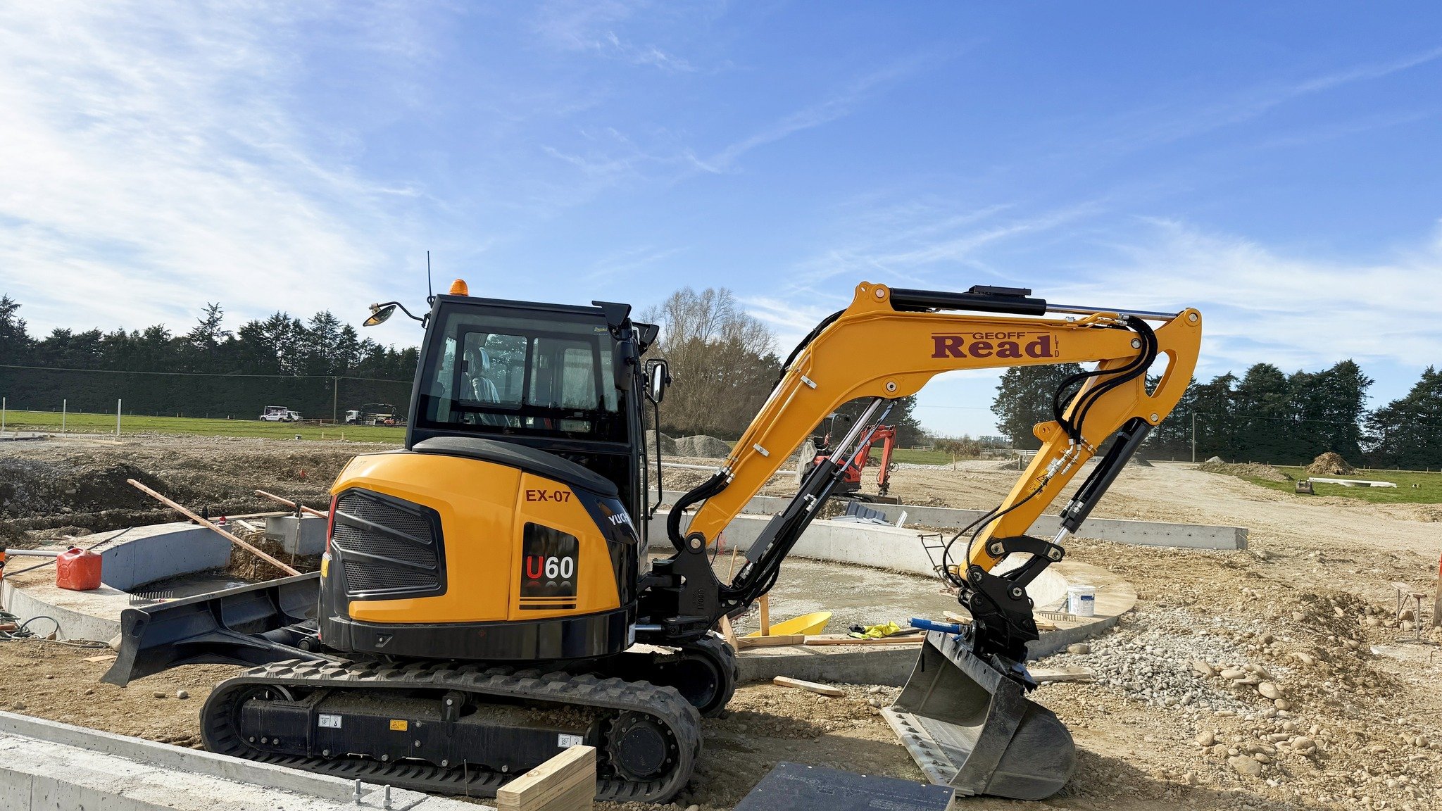Construction site with a yellow mini excavator on dirt ground, under a blue sky with some clouds, trees in the background.